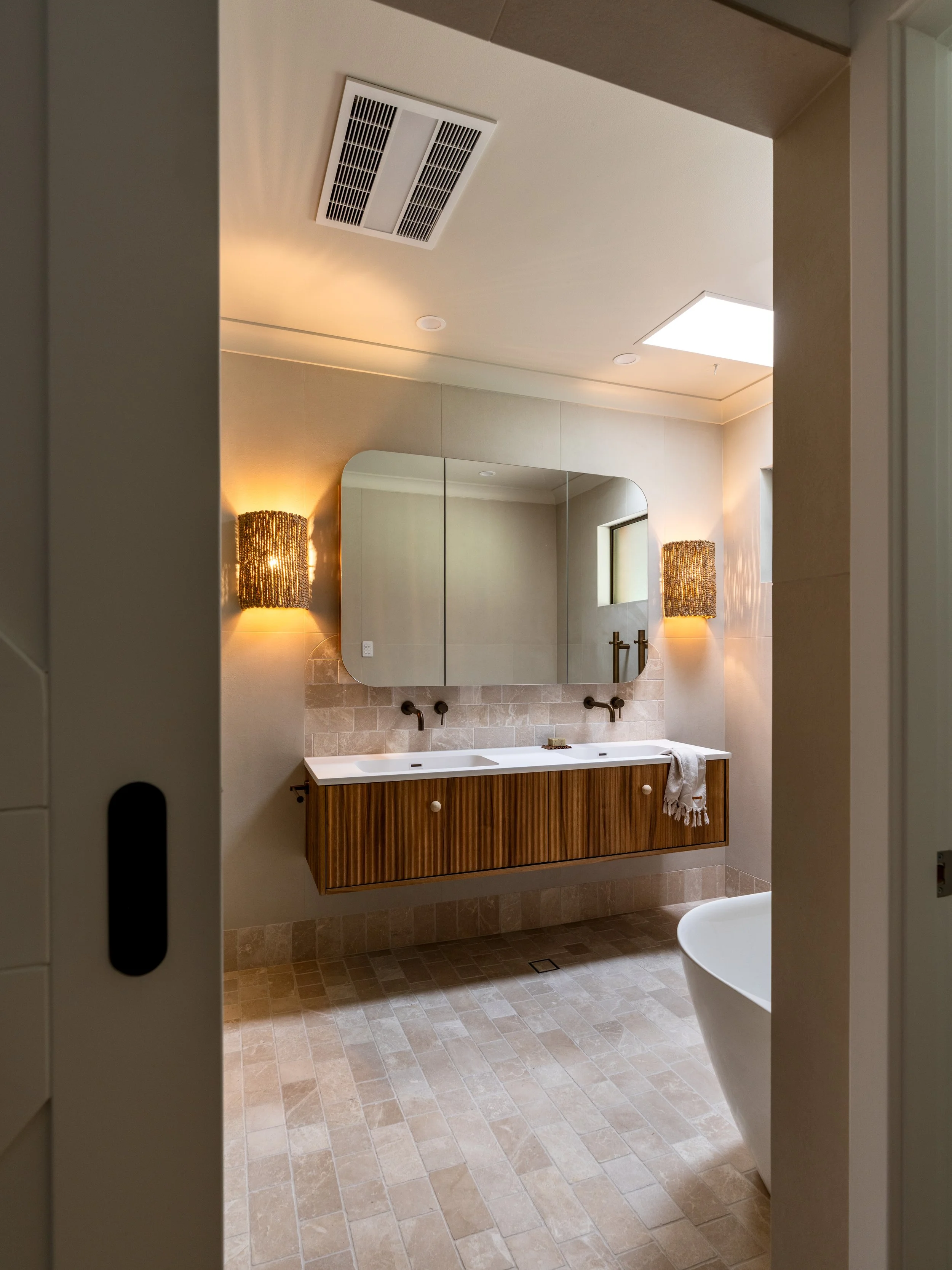 Modern bathroom with beige tile floor, wall-mounted wooden vanity with a white sink, large mirror, wall lamps, small window, and a partial view of a white freestanding bathtub.