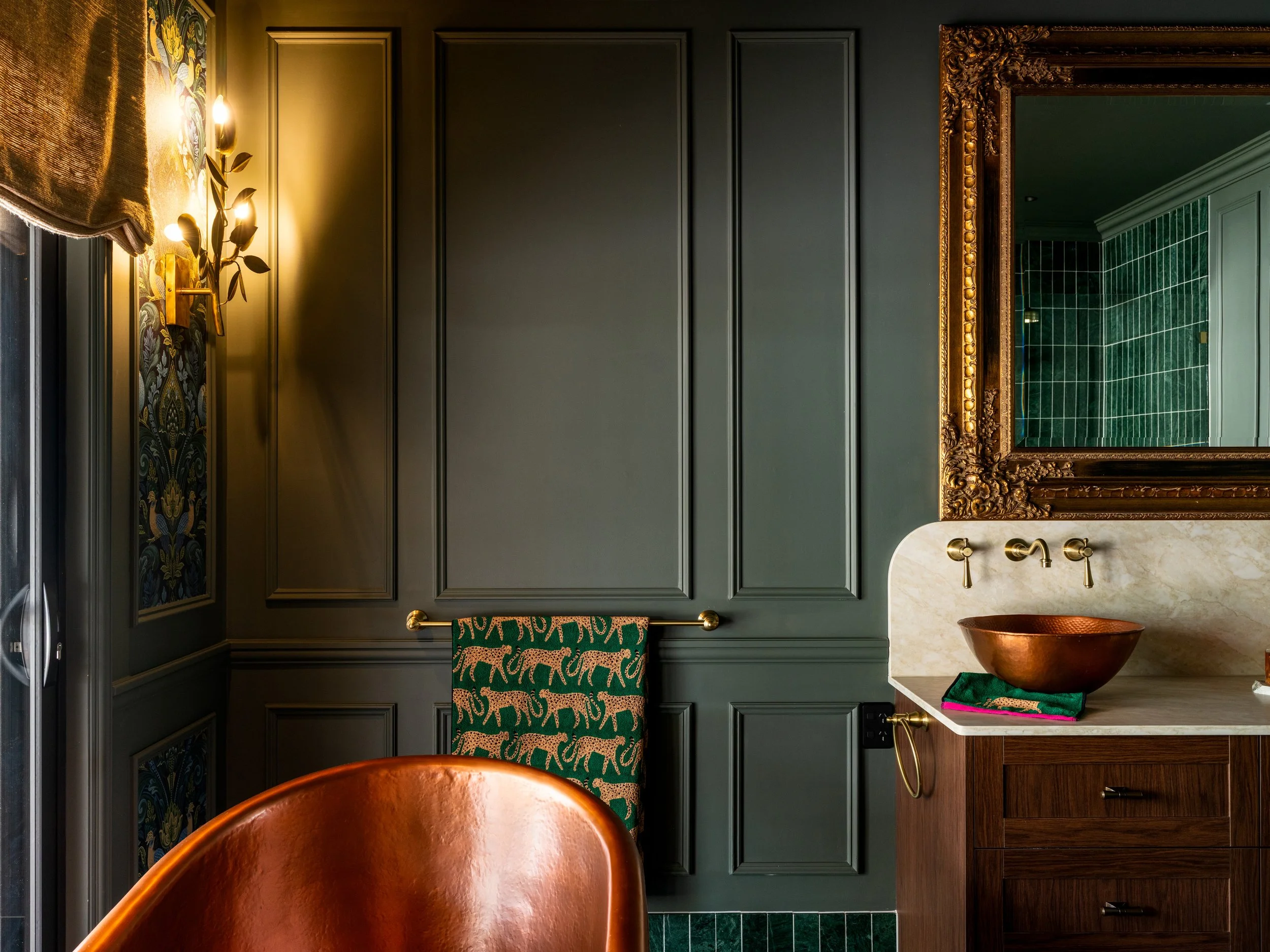 A bathroom with dark green paneled walls, a large ornate gold framed mirror, a marble countertop with a copper bowl, a wooden cabinet, and a green towel with animal patterns hanging on a gold towel rack, illuminated by wall-mounted lights.