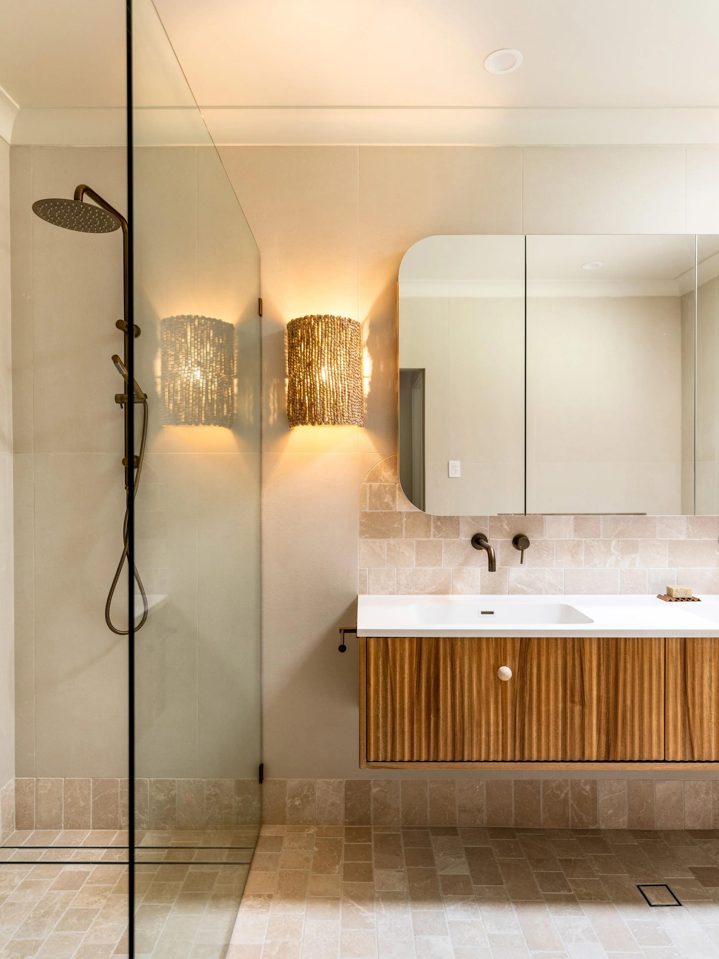 Modern bathroom with a walk-in shower on the left, featuring a brass showerhead and a hand shower. The right side has a mirrored cabinet above a white sink with a wooden cabinet below, and a decorative wall lamp with a textured shade. The floor and lower wall are tiled in beige.