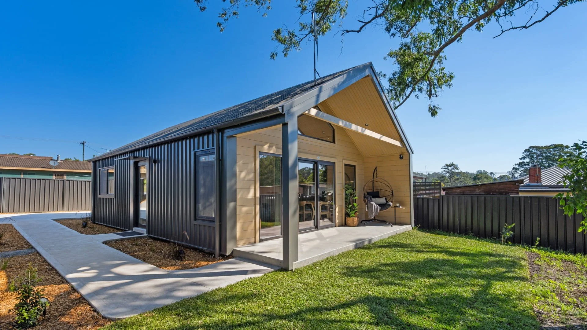 Modern house with black metal and beige siding exterior, a covered porch with a hanging chair, and a well-maintained green lawn, under a clear blue sky.