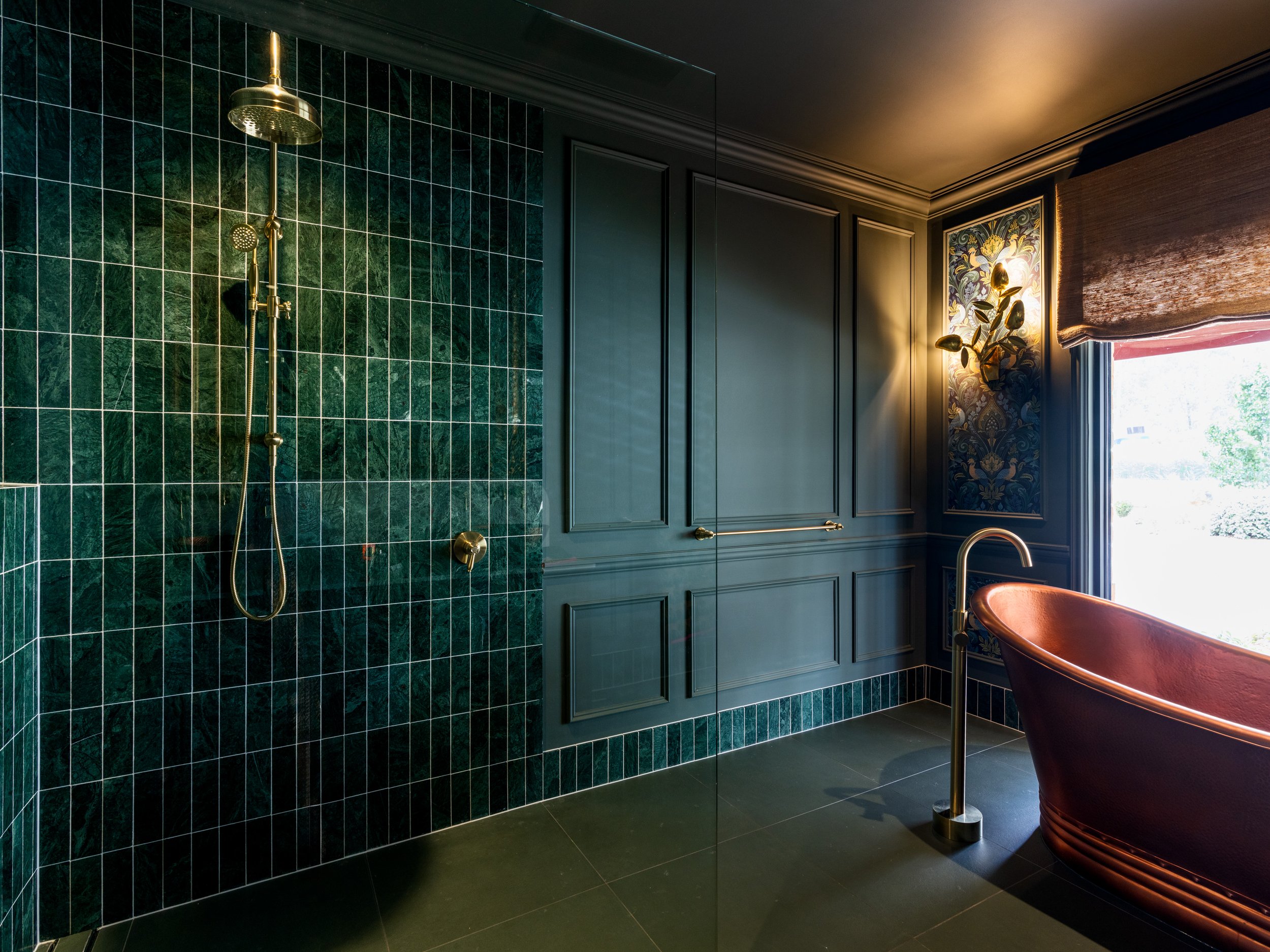A luxurious bathroom with dark green tiled shower area, gold fixtures, and a window with a scenic view and curtain, featuring a red bathtub and a decorative wall with floral wallpaper and a gold wall sconce.