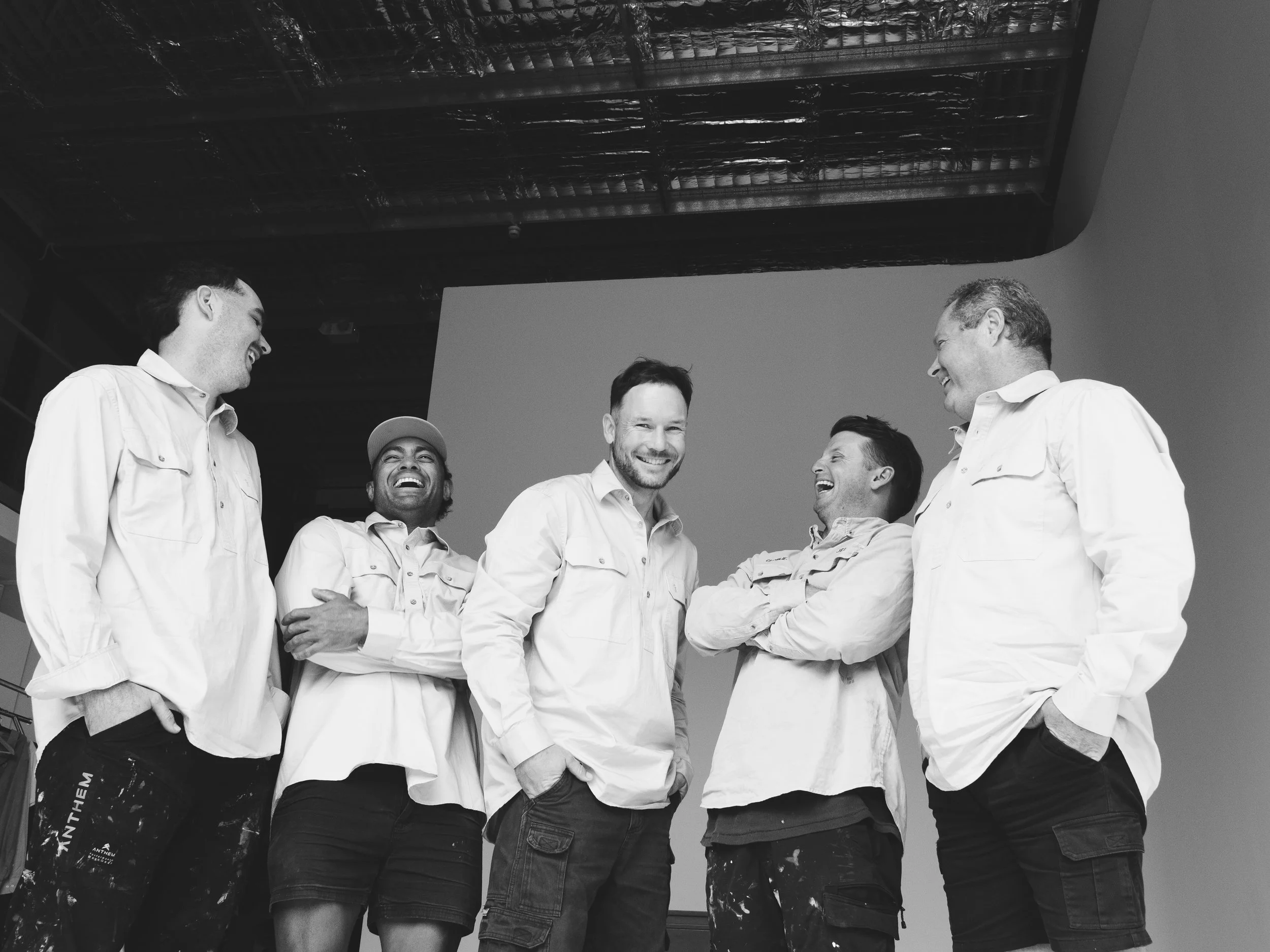 Six men wearing white shirts are standing in a circle, smiling and laughing together, in an indoor setting with a beam ceiling.
