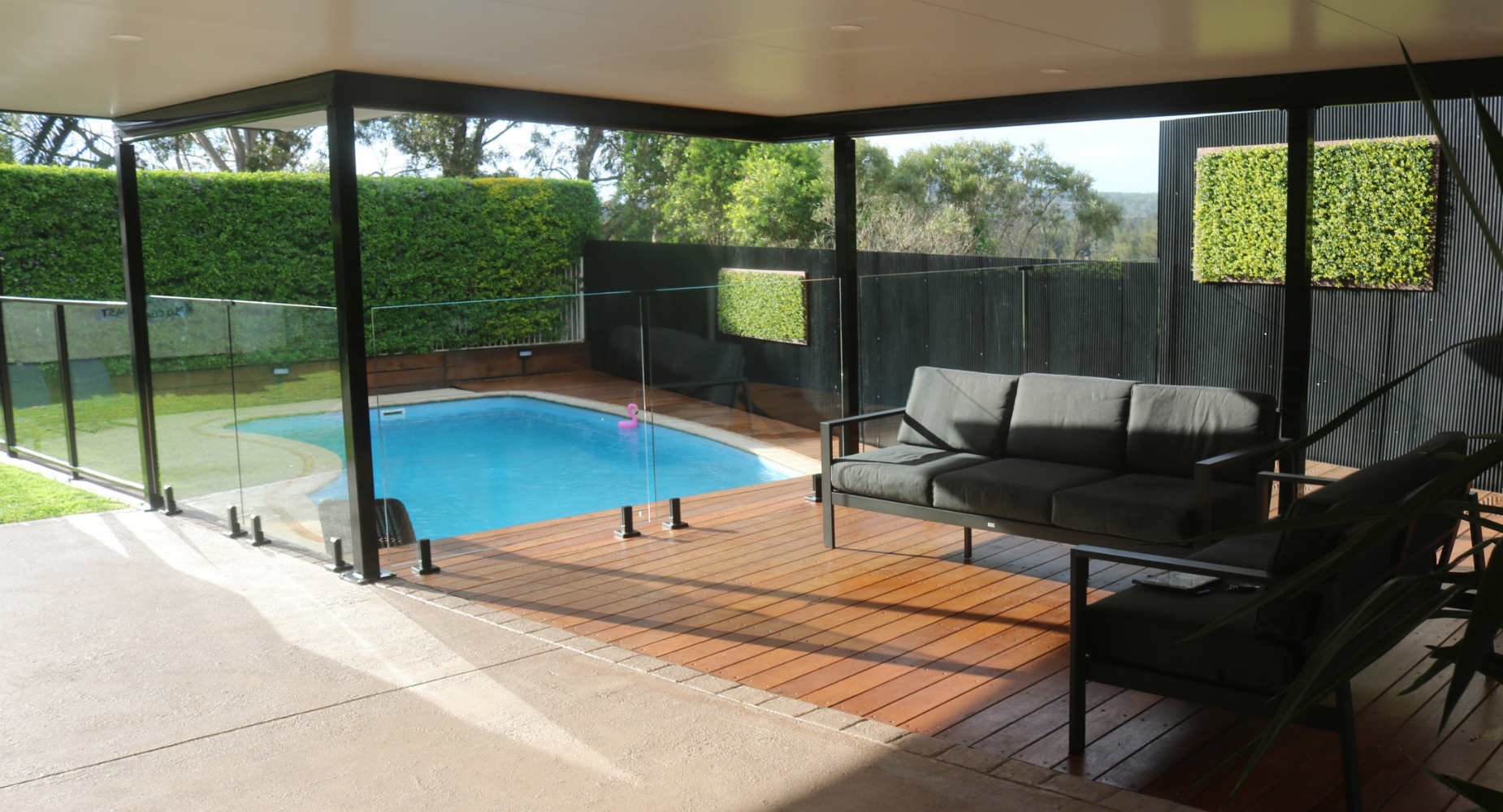 Patio with a glass fence, featuring a swimming pool with a pink flamingo float, and outdoor seating including a black sofa, in a backyard surrounded by greenery.