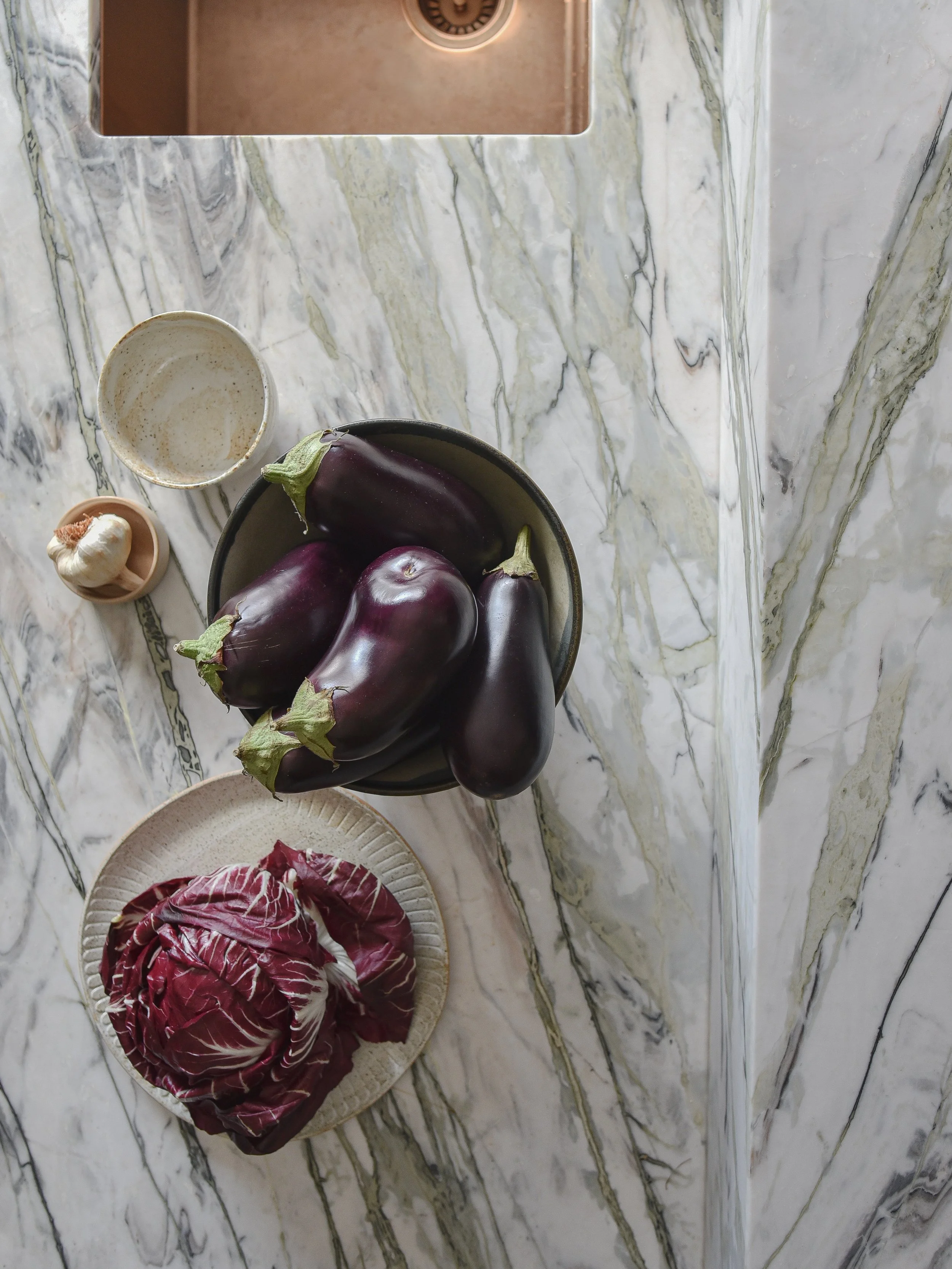A marble kitchen countertop with a bowl of eggplant, a bowl of garlic, a bowl with cream or sauce, and two plates of radicchio.