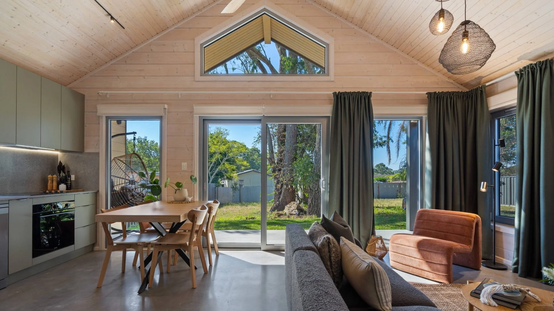Living room with large sliding glass doors, green curtains, wooden ceiling with pendant lights, sofa, orange armchair, dining table with chairs, and view of backyard with trees and fence.