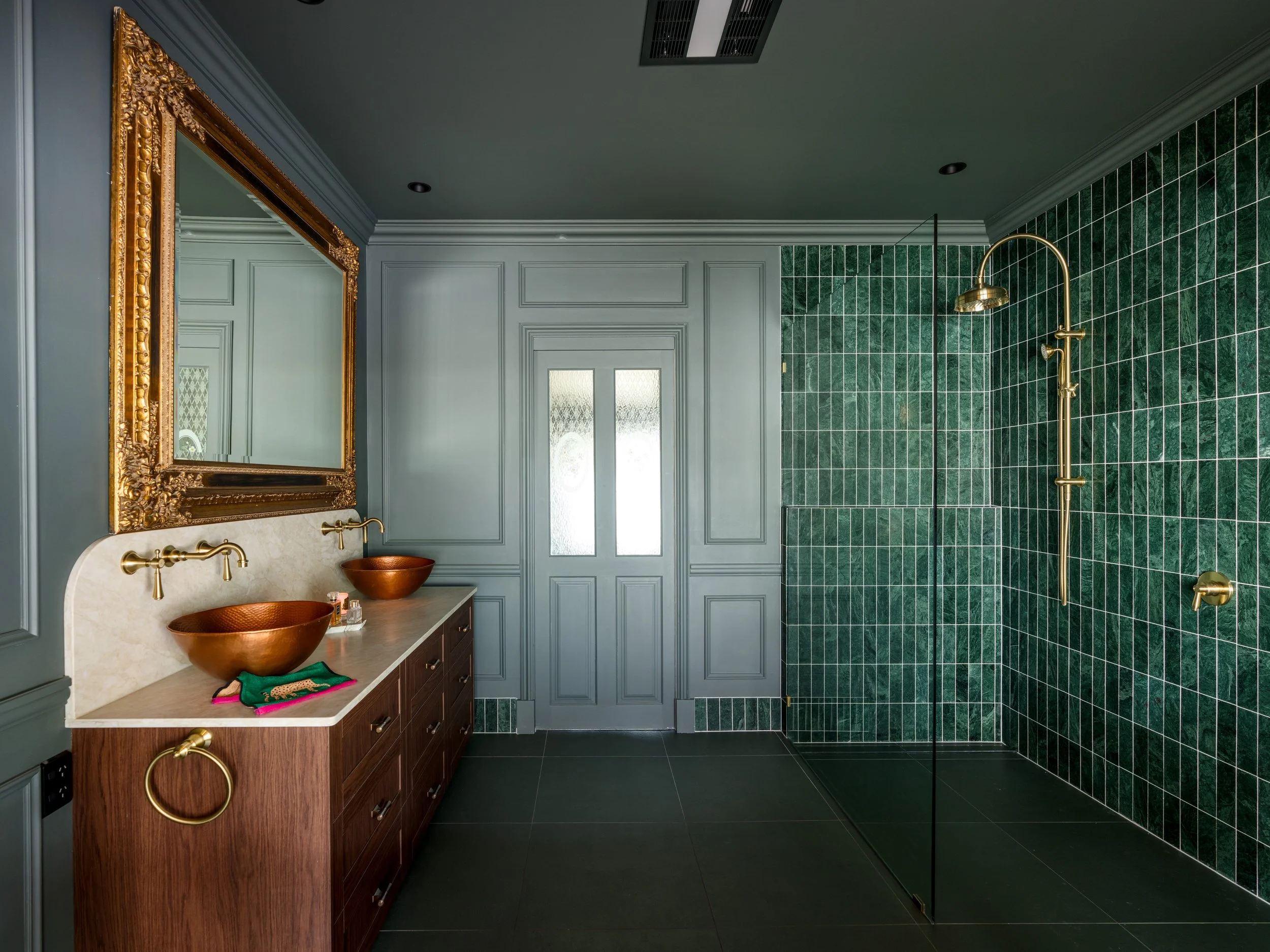 A bathroom featuring a gold-framed mirror, green tiled shower area with a gold showerhead, and a double vanity with copper vessel sinks and a wooden cabinet.