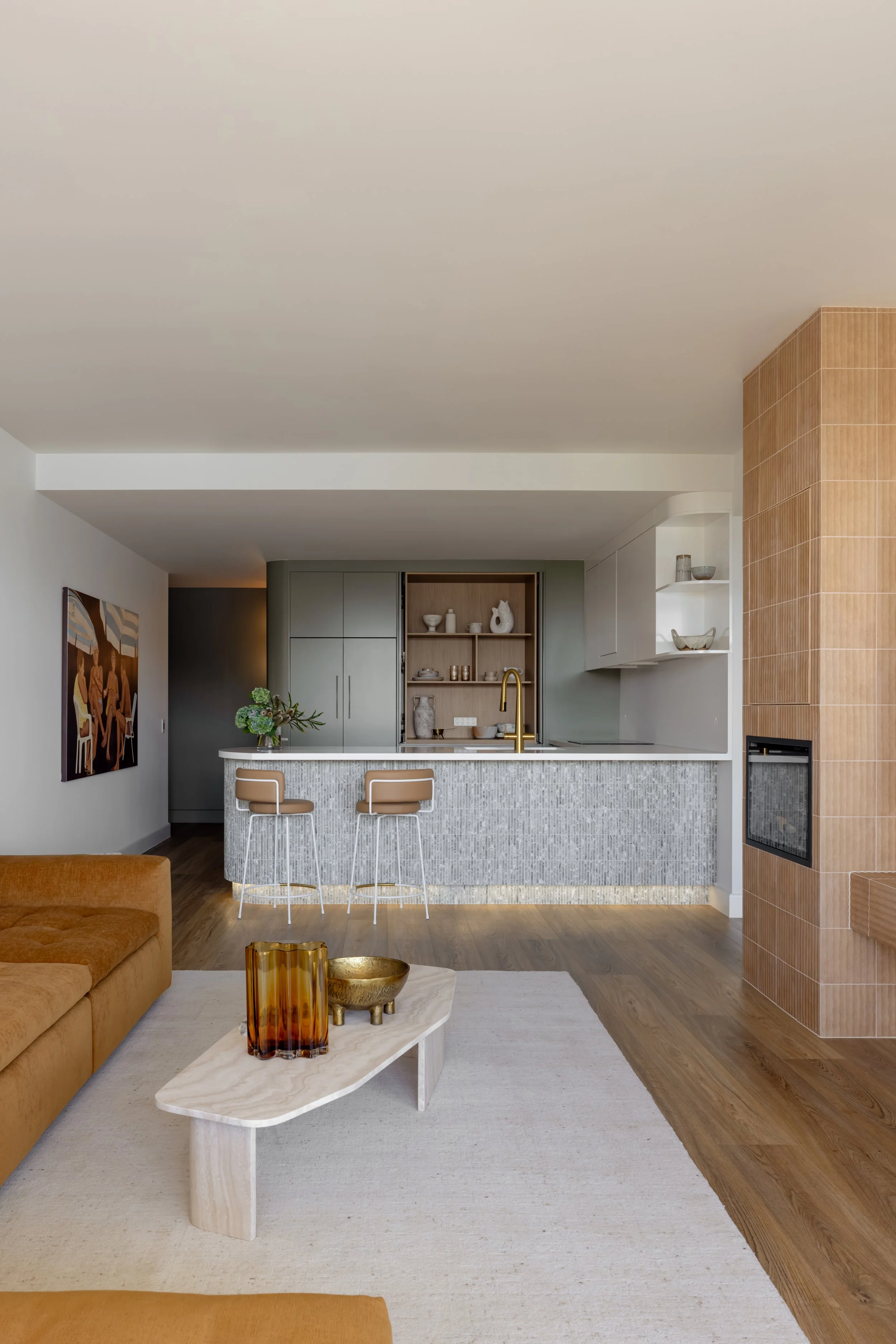 Interior view of a modern living room with a kitchen counter, two barstools, a sofa, a coffee table with decorative vases, a painting on the wall, and a combination of wood, white, and gray finishes.