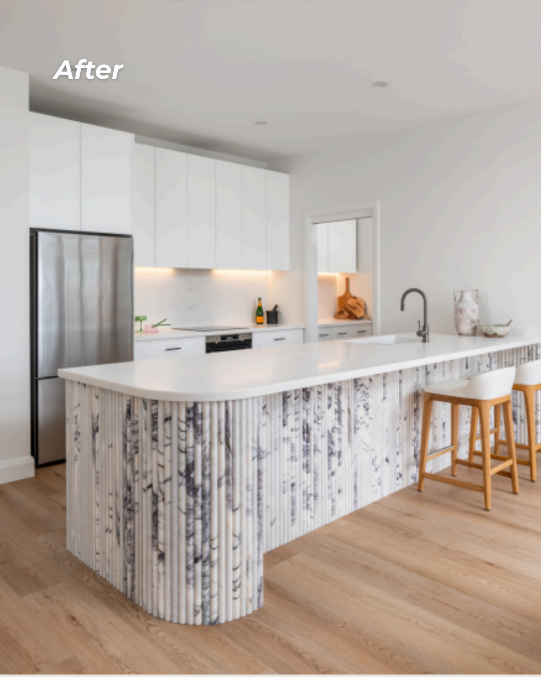 Modern kitchen with a marble-topped island featuring a ribbed, white and gray marble design on the base, white cabinetry, and wooden barstools, with the word 'After' in white text in the upper left corner.