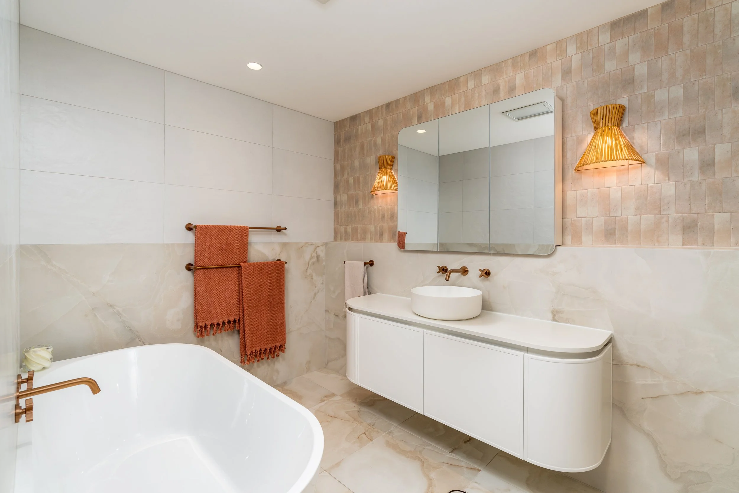 A modern bathroom with a white bathtub, a white vanity with a vessel sink, a large mirror, and textured beige and white tiled walls, accented by two wooden wall sconces and orange towels.