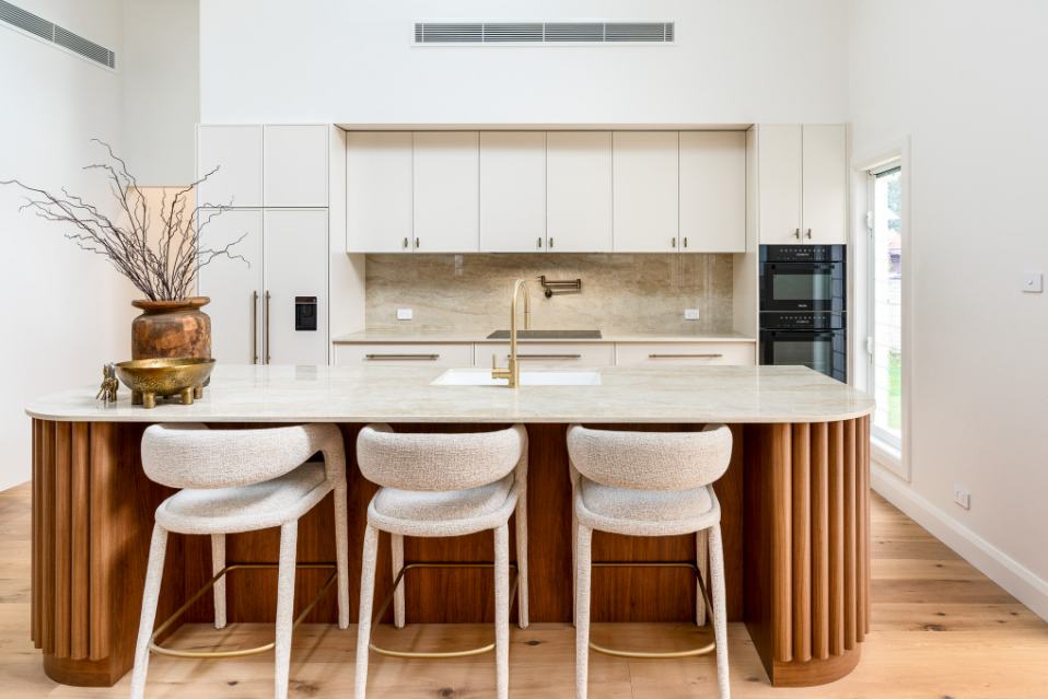 Modern kitchen with white cabinets, a large marble island with wooden paneling, three beige bar stools, and minimal decor.