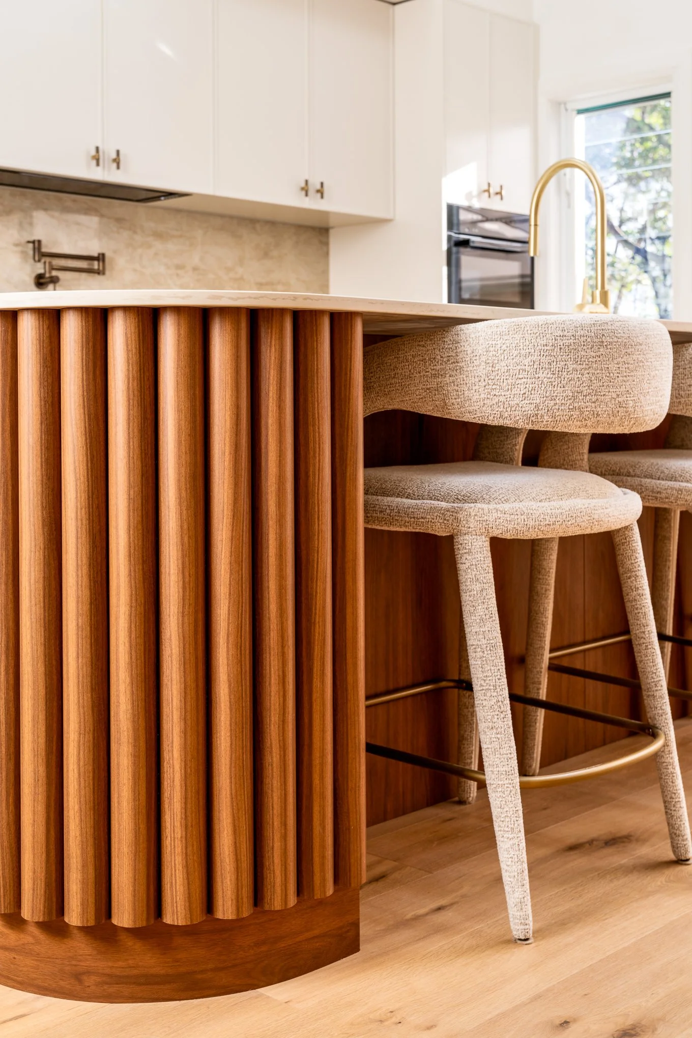 Close-up of a modern kitchen island with a wood slat design, beige upholstered bar stool, marble countertop, white cabinetry, gold faucet, and a window with a view of trees.