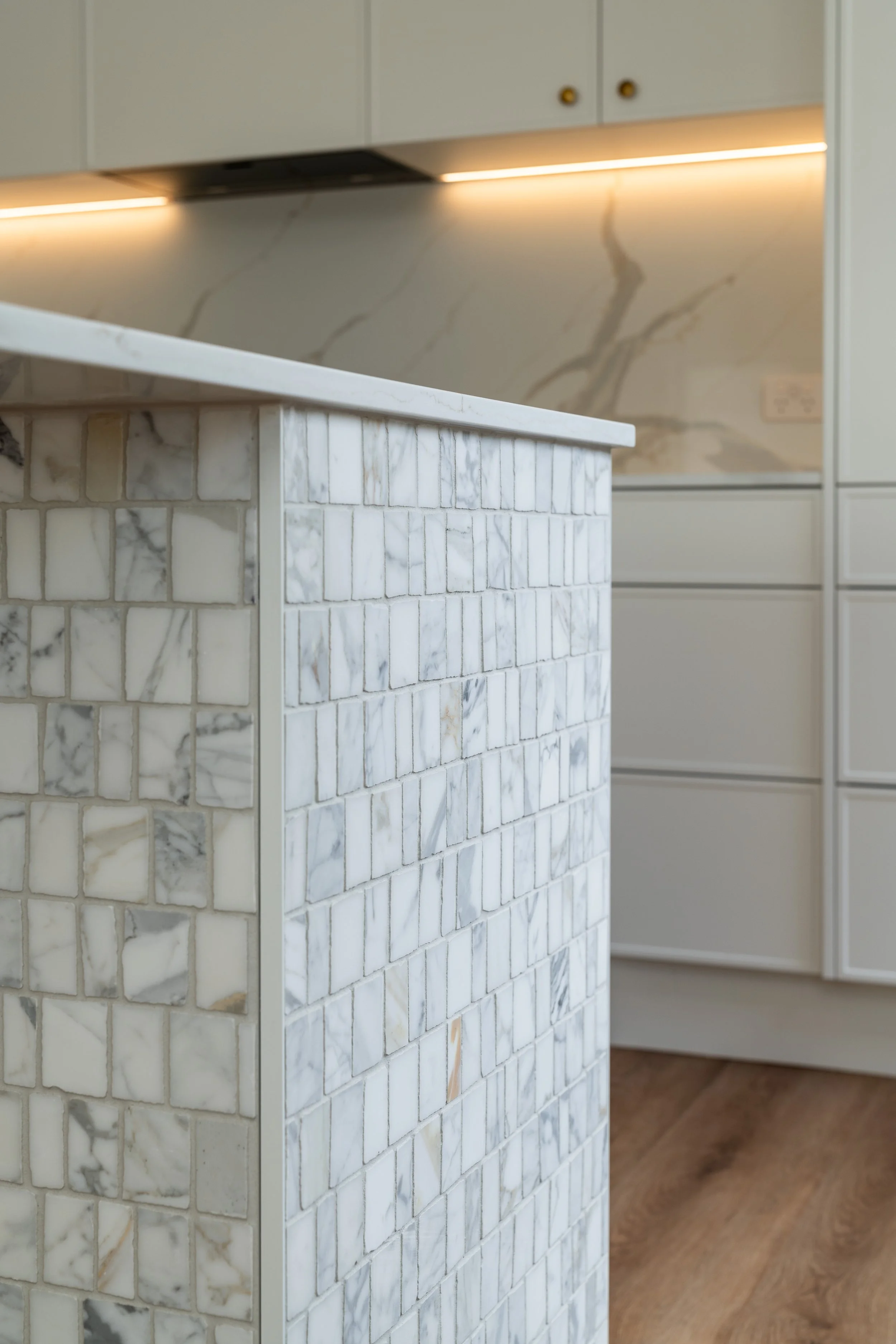 Close-up of a kitchen island with a marble tile surface and a marble tile front, with a marble backsplash and upper cabinets in the background.