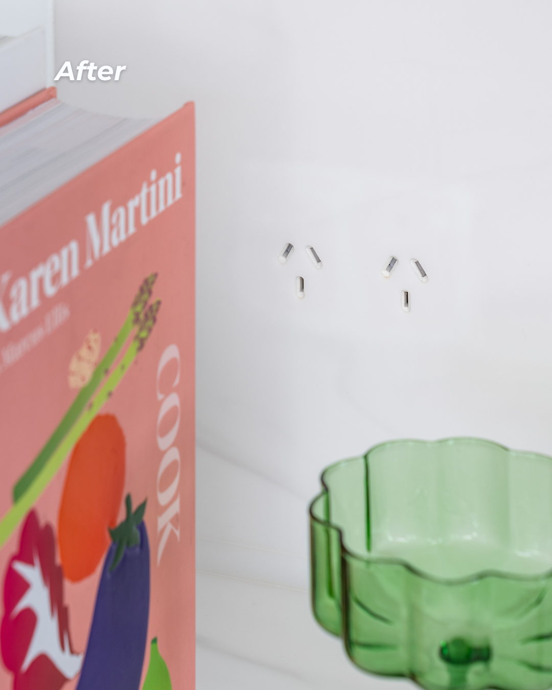 A pink book titled Karen Martini with fruit illustrations partially visible on the left, a light green glass bowl on the right, and a white wall with small metallic hooks in the background.