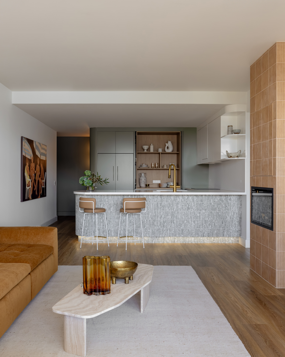 Modern open-concept living room with a kitchen featuring a marble island, beige barstools, and cabinetry with decorative pottery, a gold faucet, and a built-in fireplace with terracotta tiles.