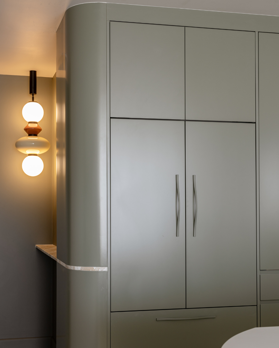 A modern, minimalist kitchen cabinet with two elongated handles and a curved wall with a light gray finish, next to a wall with a decorative hanging light fixture.