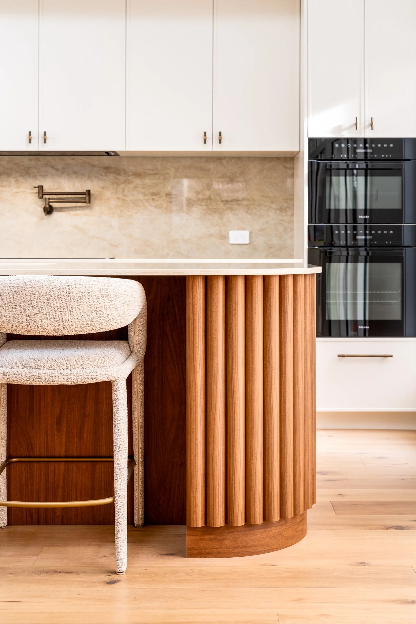 A modern kitchen with white upper cabinets, a black oven, beige marble backsplash, wooden cabinetry, a beige upholstered chair, and a wooden floor.
