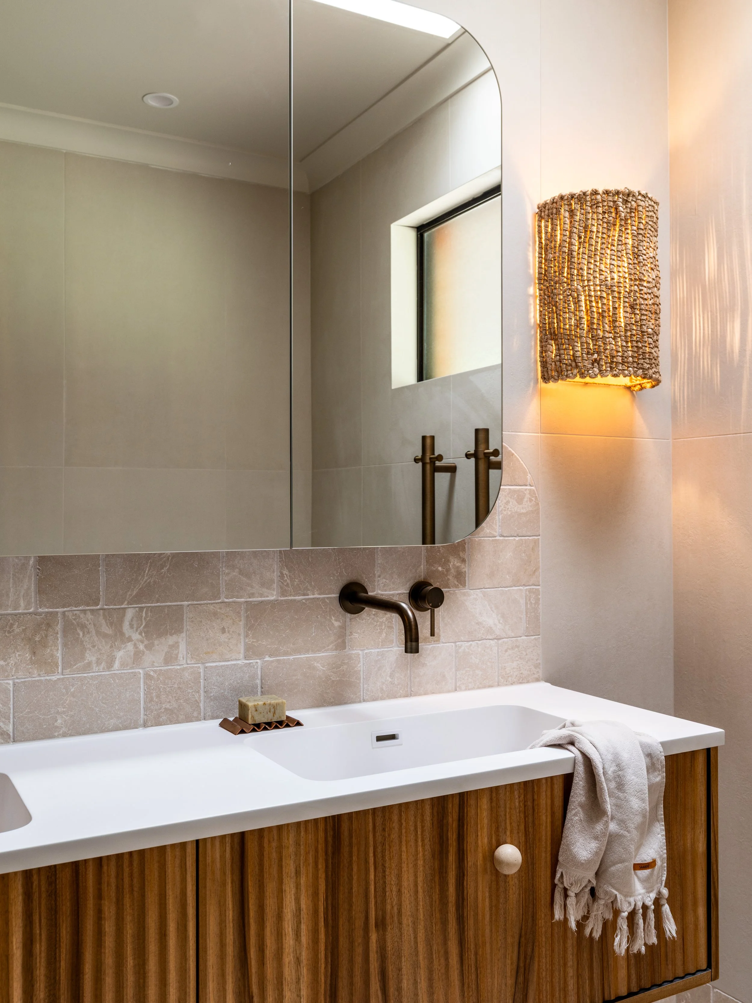 A bathroom vanity with a large mirror, a beige tile wall, a white sink, a soap bar, a towel hanging on a towel rack, and a decorative wall sconce with warm lighting.