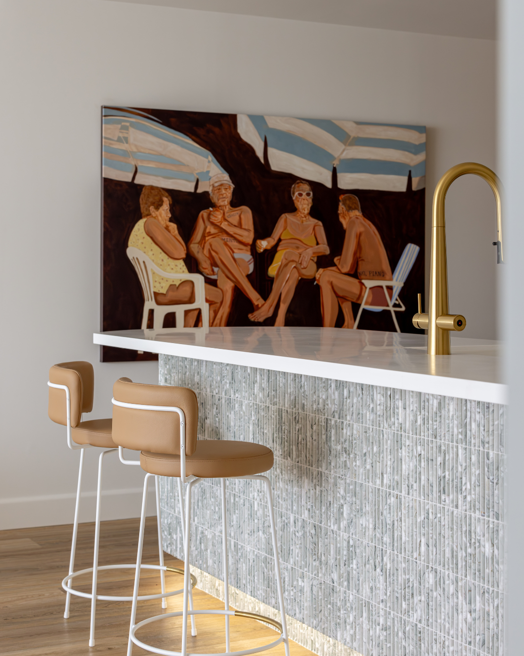 A modern kitchen with a white counter, gold faucet, and beige bar stools, featuring a large painting of four elderly people sitting under striped umbrellas.
