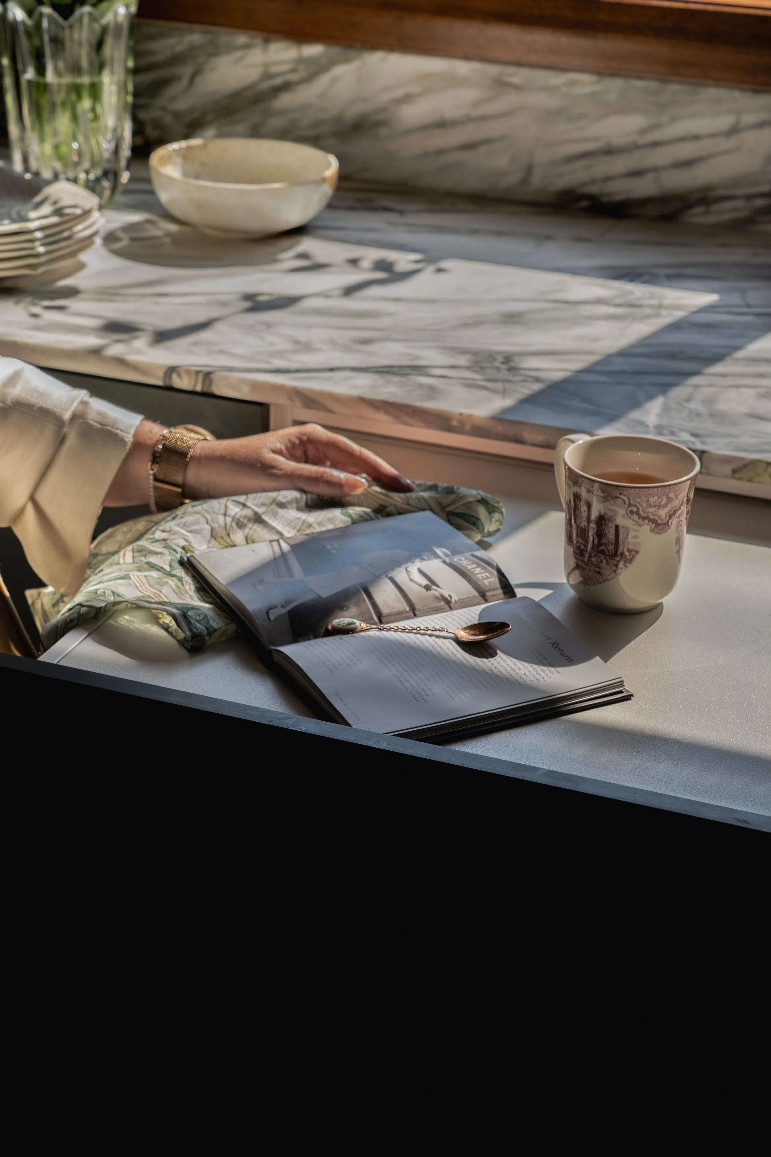 A person wearing a white shirt and gold watch sitting at a table with a coffee mug, an open magazine, and a folded cloth napkin. There are plates, bowls, and glasses in the background on a marble countertop.