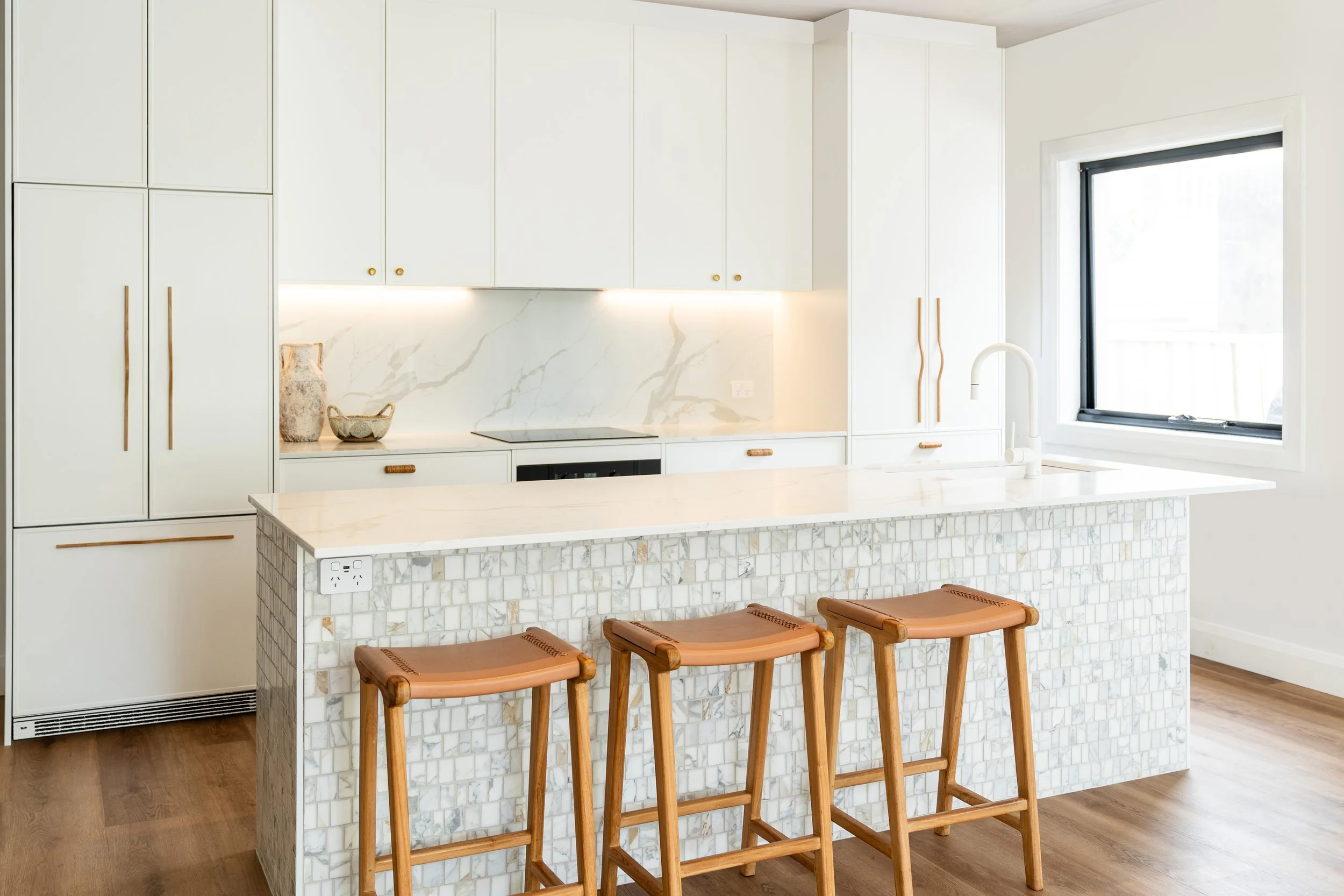 Modern kitchen with white cabinetry, marble backsplash, marble kitchen island, wooden bar stools, and large window with black trim.