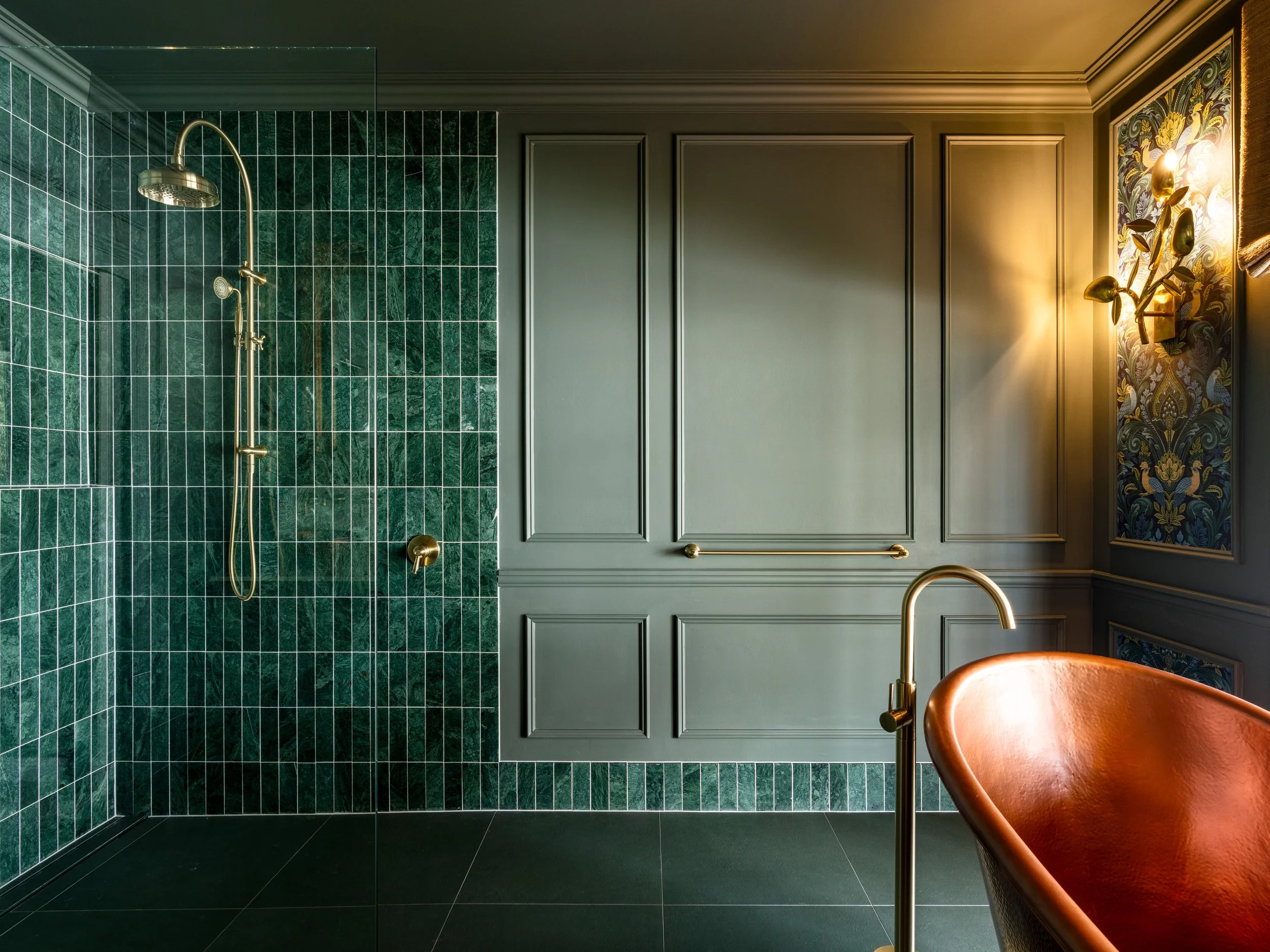 A modern bathroom featuring a green tiled shower area with brass fixtures, grey paneled walls, and a decorative floral wallpaper on one side, with a warm-toned orange chair visible in the foreground.