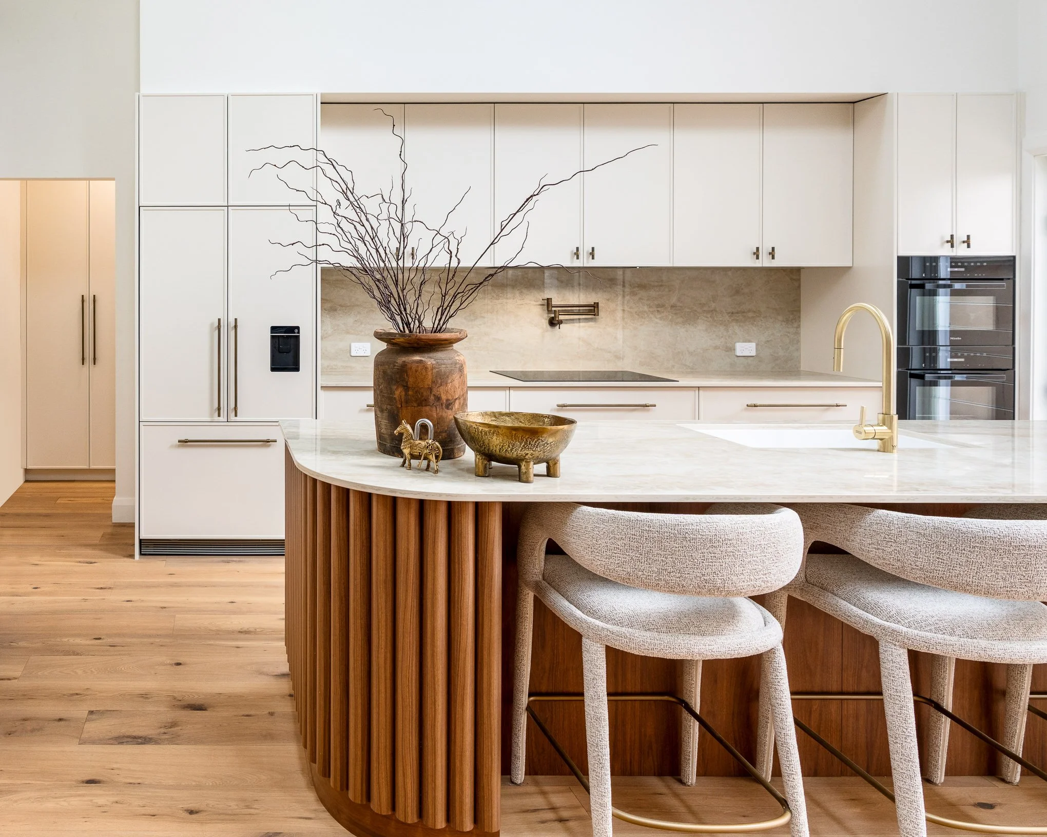 Modern kitchen with white cabinets, a white marble island with wooden paneling, decorative bowls, dried branches in a large vase, and a gold faucet.