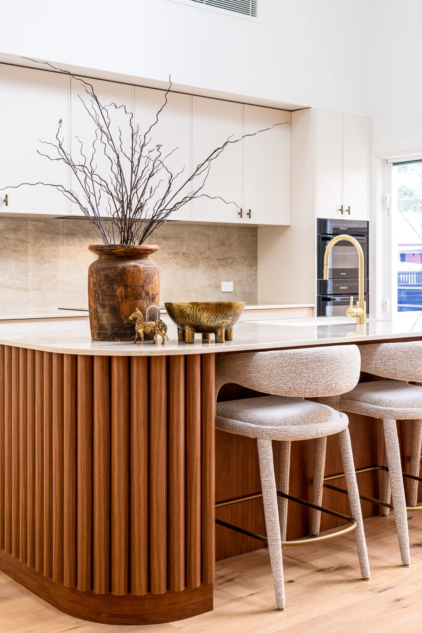Modern kitchen with white cabinetry, a wooden kitchen island with vertical slats, a gold faucet, and barstools with light-colored fabric seats. Decor includes a large wooden vase with branches, a small metallic horse figurine, and a decorative bowl.