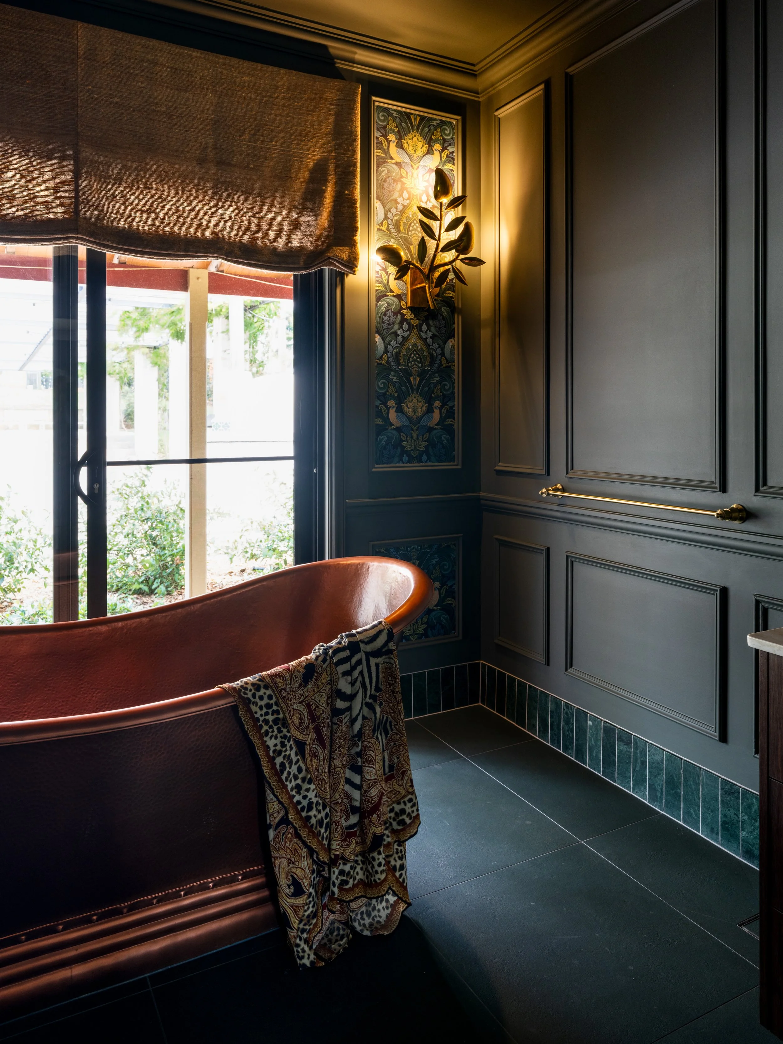 Interior of a room with dark blue paneled walls, a window with a brown curtain, a copper bathtub with patterned towels hanging over its side, decorative wallpaper behind a wall sconce, and tiled flooring.
