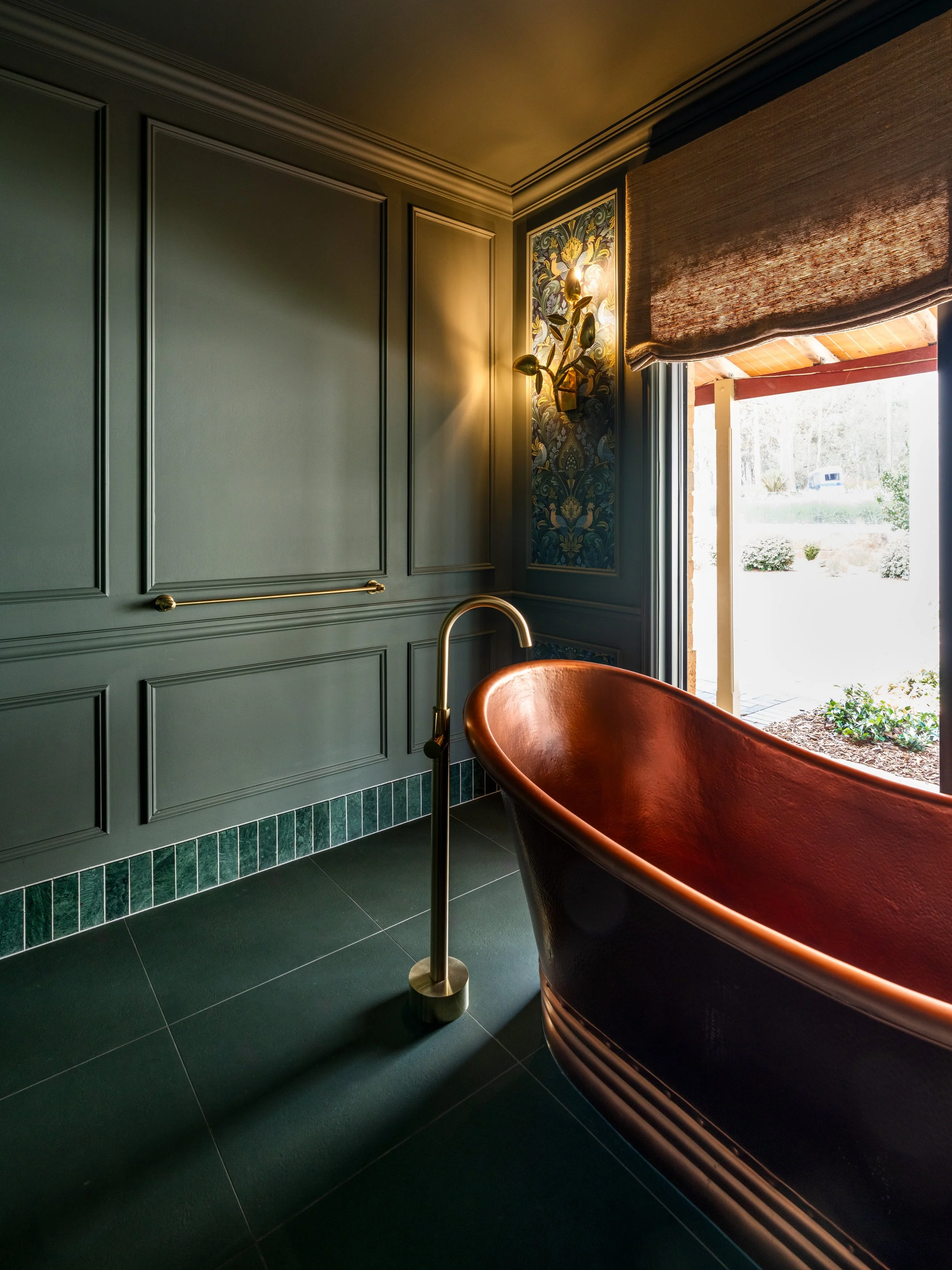 A bathroom with dark green wall paneling, a copper bathtub, black tiled floor, and a window with a woven shade. Sunlight coming through the window, outside greenery visible.
