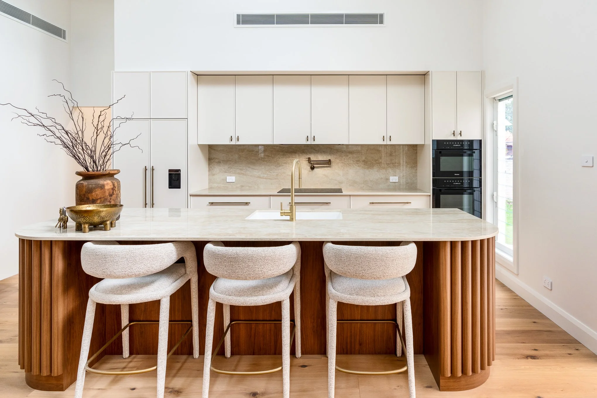 Modern kitchen with white cabinets, marble island with a gold faucet, and four beige upholstered bar stools. Decor includes a large vase with branches and a gold bowl on the island. Light wooden flooring and a window are visible.