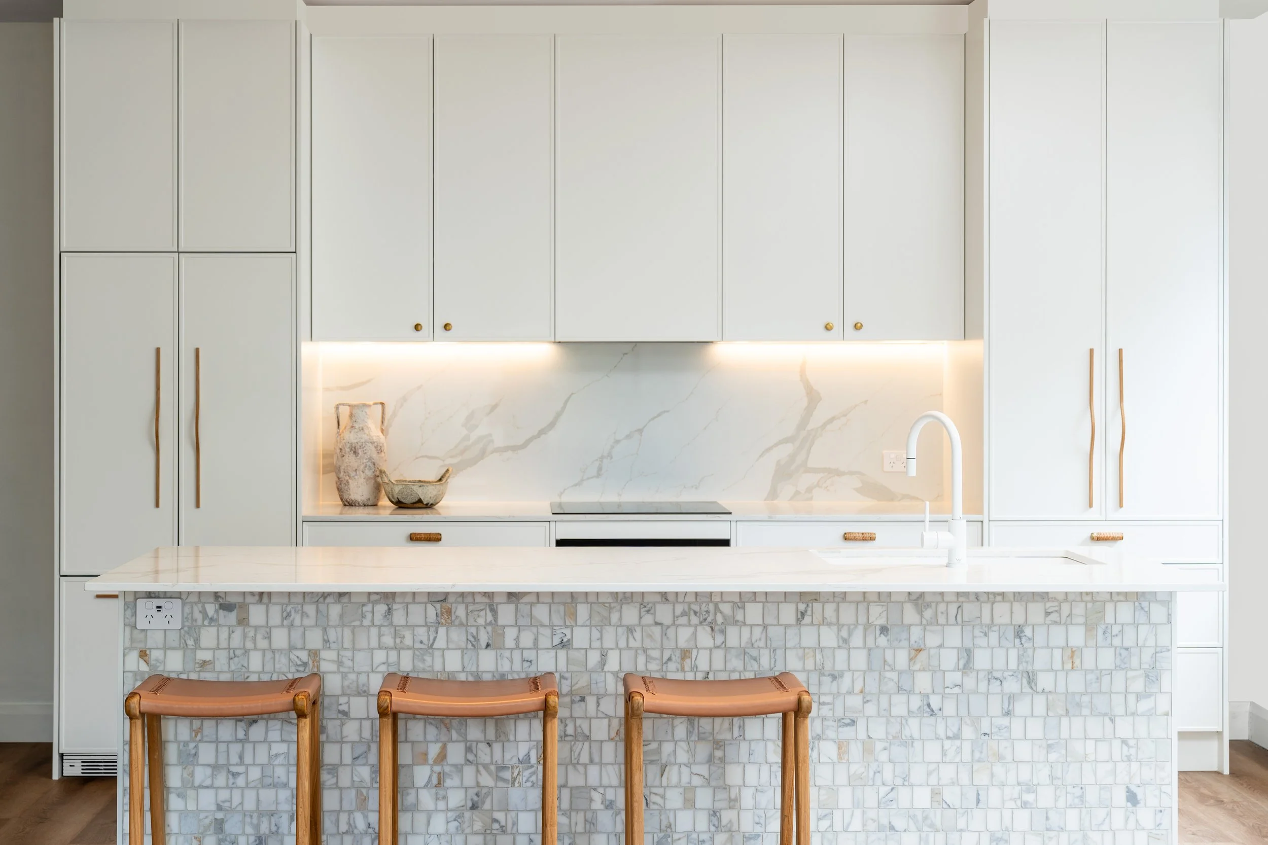 Modern kitchen with white cabinetry, marble backsplash, and a marble kitchen island with a built-in cooktop. There are three wooden stools at the island, decorative vases on the countertop, and a sink with a white faucet. A power outlet is visible on