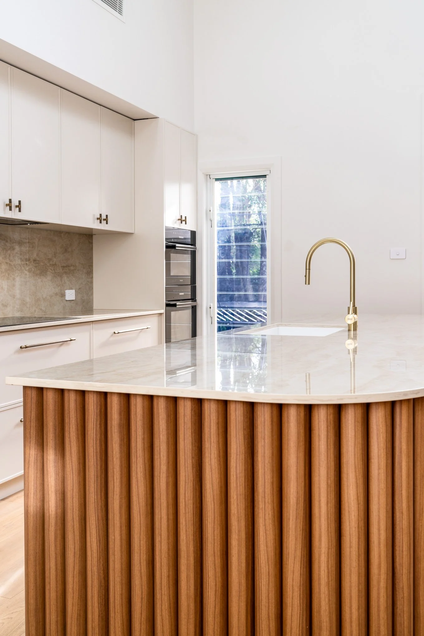 Modern kitchen with a marble island, gold faucet, white cabinets, double oven, and a sliding glass door leading outside.