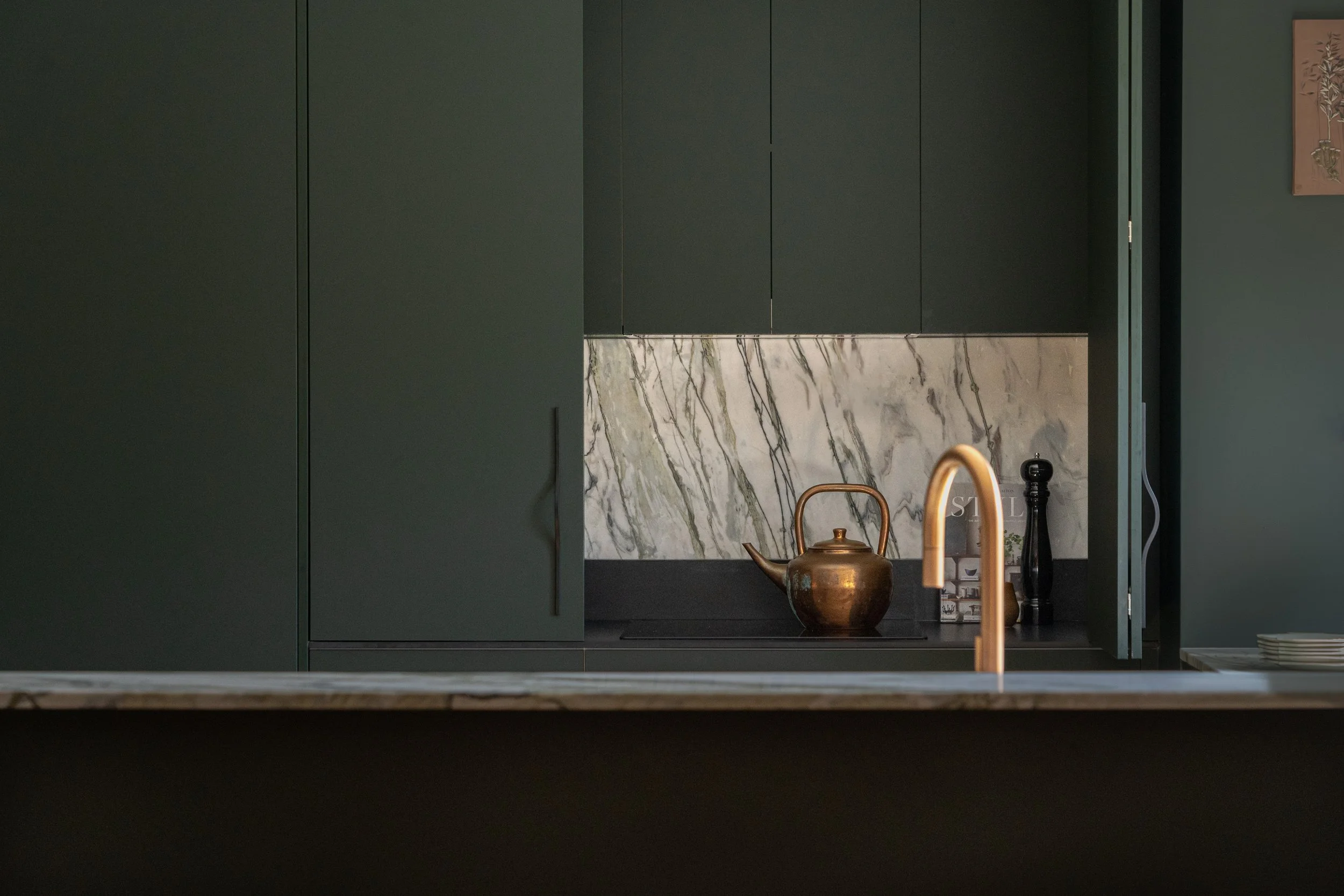 A kitchen scene with dark green cabinets, a marble backsplash, a gold teapot, a black pepper grinder, and a gold-colored faucet.