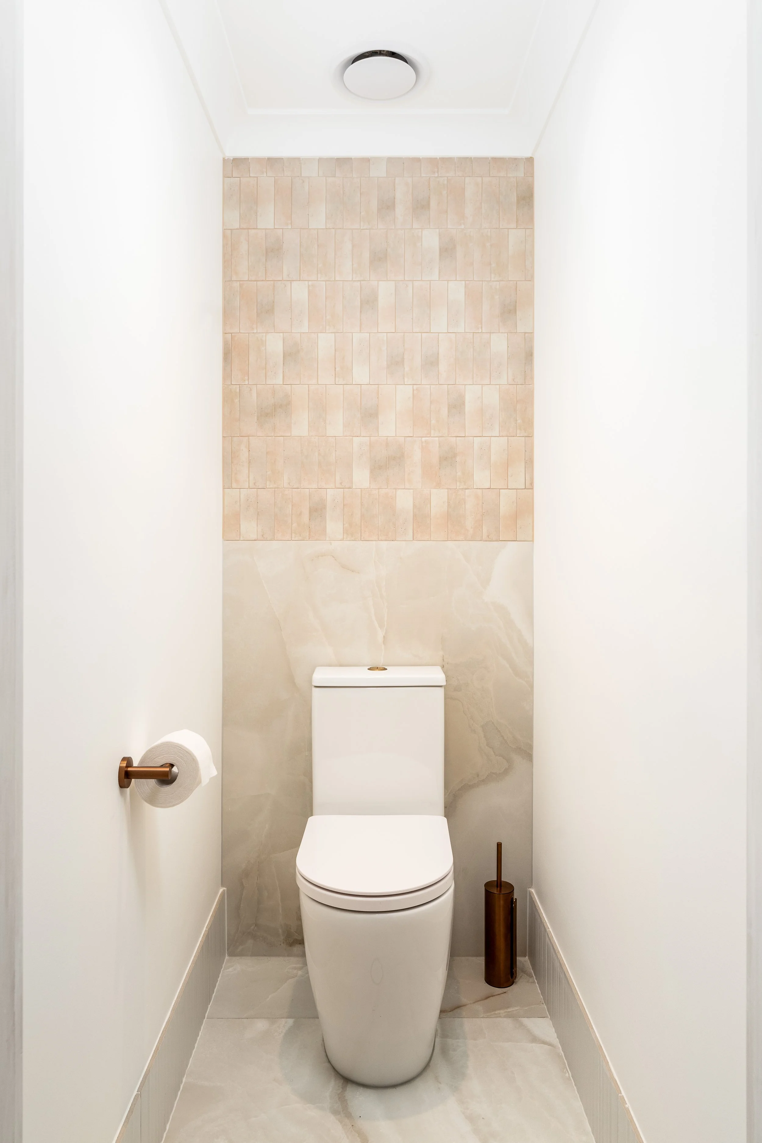 Small bathroom with modern toilet, beige tile wall behind it, white walls, and marble floor, with a brown toilet brush holder and a white toilet paper holder with a roll of toilet paper.
