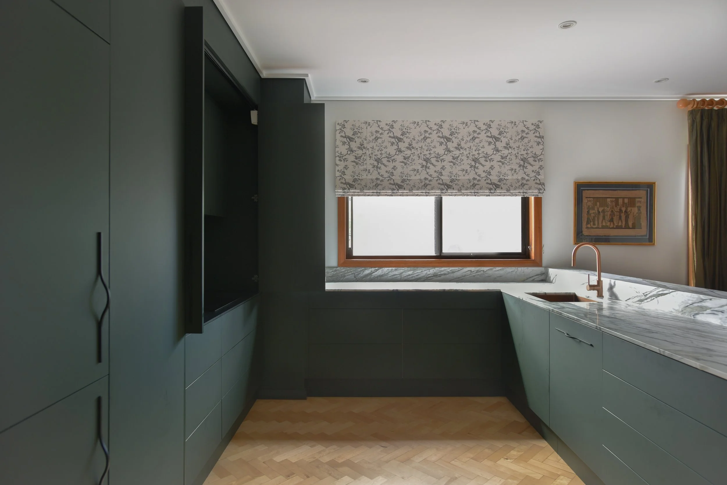 Kitchen with dark green cabinets, a window with a floral Roman shade, a framed artwork on the wall, and a marble countertop with a copper faucet.