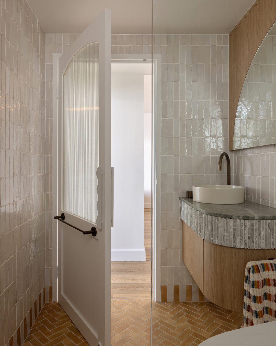 A small bathroom with beige tiled walls and a wooden floor. It features a curved mirror, a modern sink with a dark faucet, and a vanity with a textured front. An open door reveals a glimpse of the adjacent room.