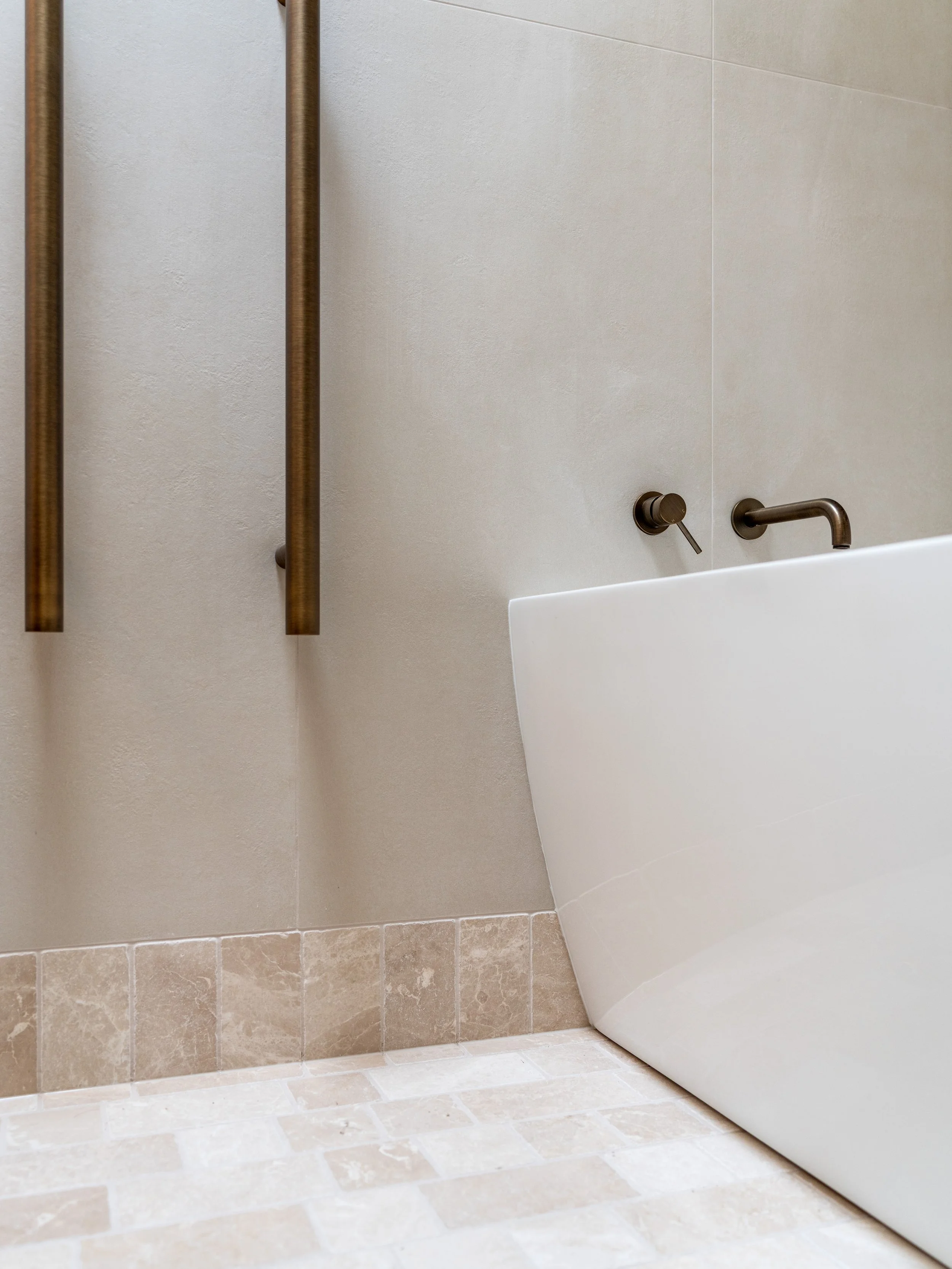 Close-up of a bathroom corner showing a white bathtub, beige floor tiles, and beige wall tiles. There are two metallic fixtures, one for water and the other possibly a control knob, mounted on the wall above the bathtub. Two wooden towel bars are ins