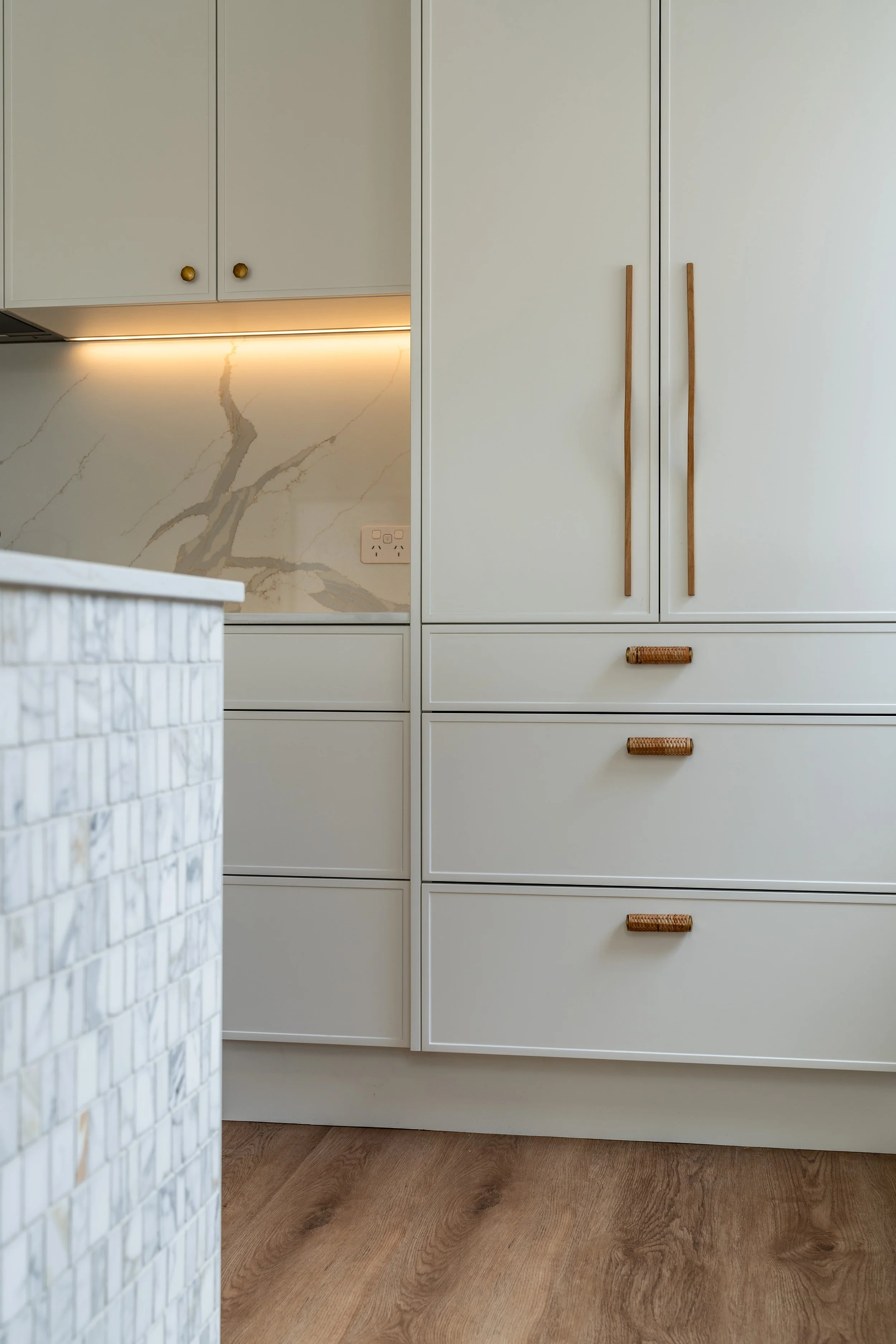 Close-up of white kitchen cabinets with minimalist wooden handles, marble backsplash with gold veining, and a power outlet. Part of a kitchen island with a textured white surface is visible in the foreground, and the floor is wood.
