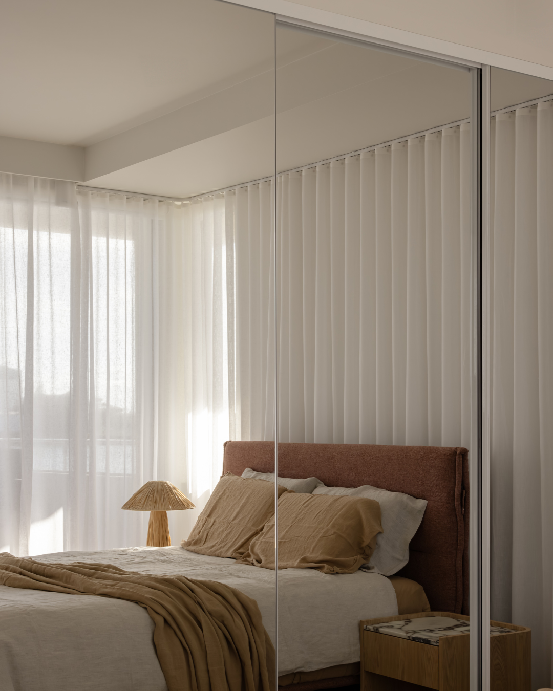 A bedroom with a bed, beige and white bedding, a brown headboard, a wooden bedside table, a lamp with a straw shade, and white curtains covering large windows.