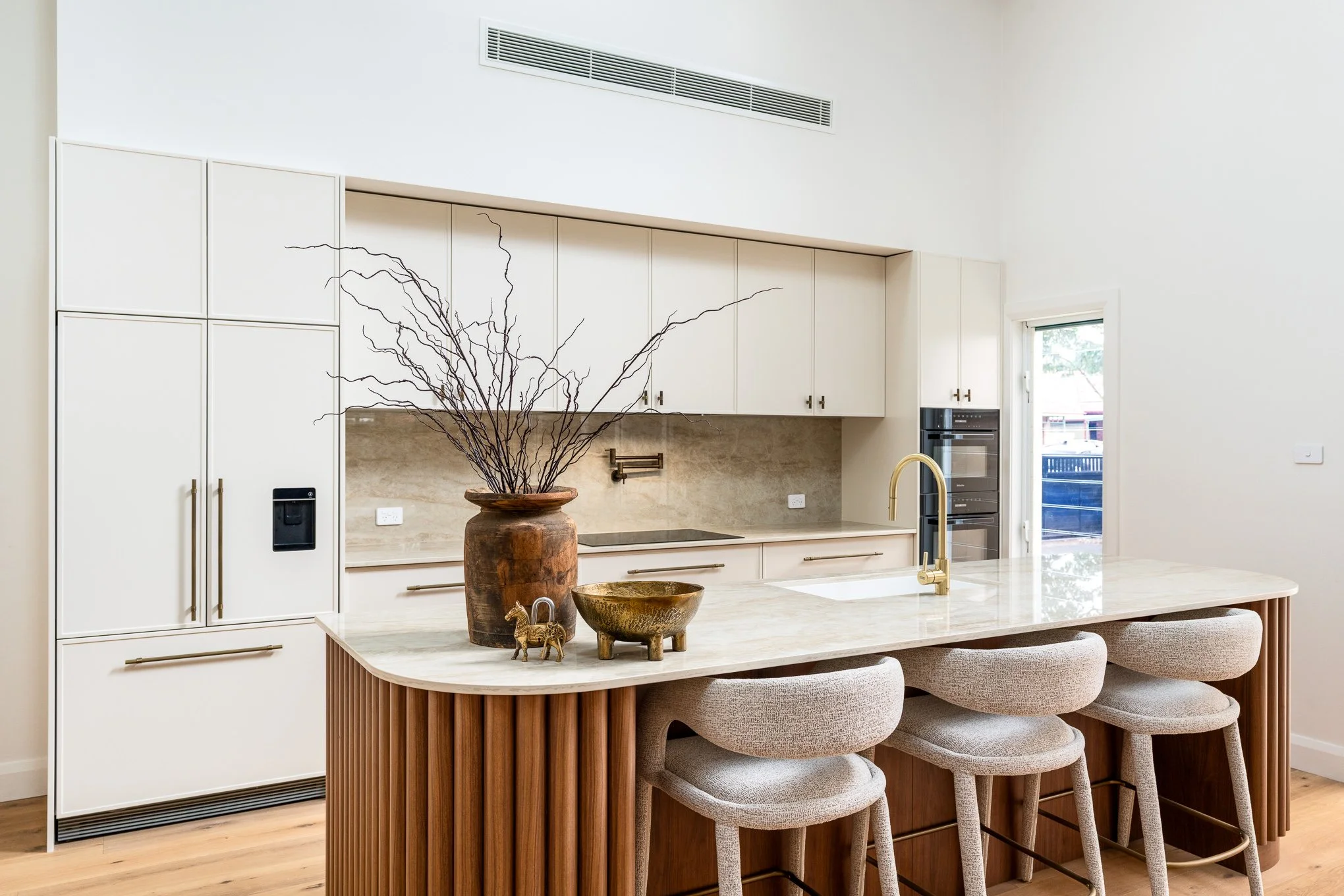 Modern kitchen with white cabinets, a marble island with beige stools, and decorative items like a brown vase and gold accents.