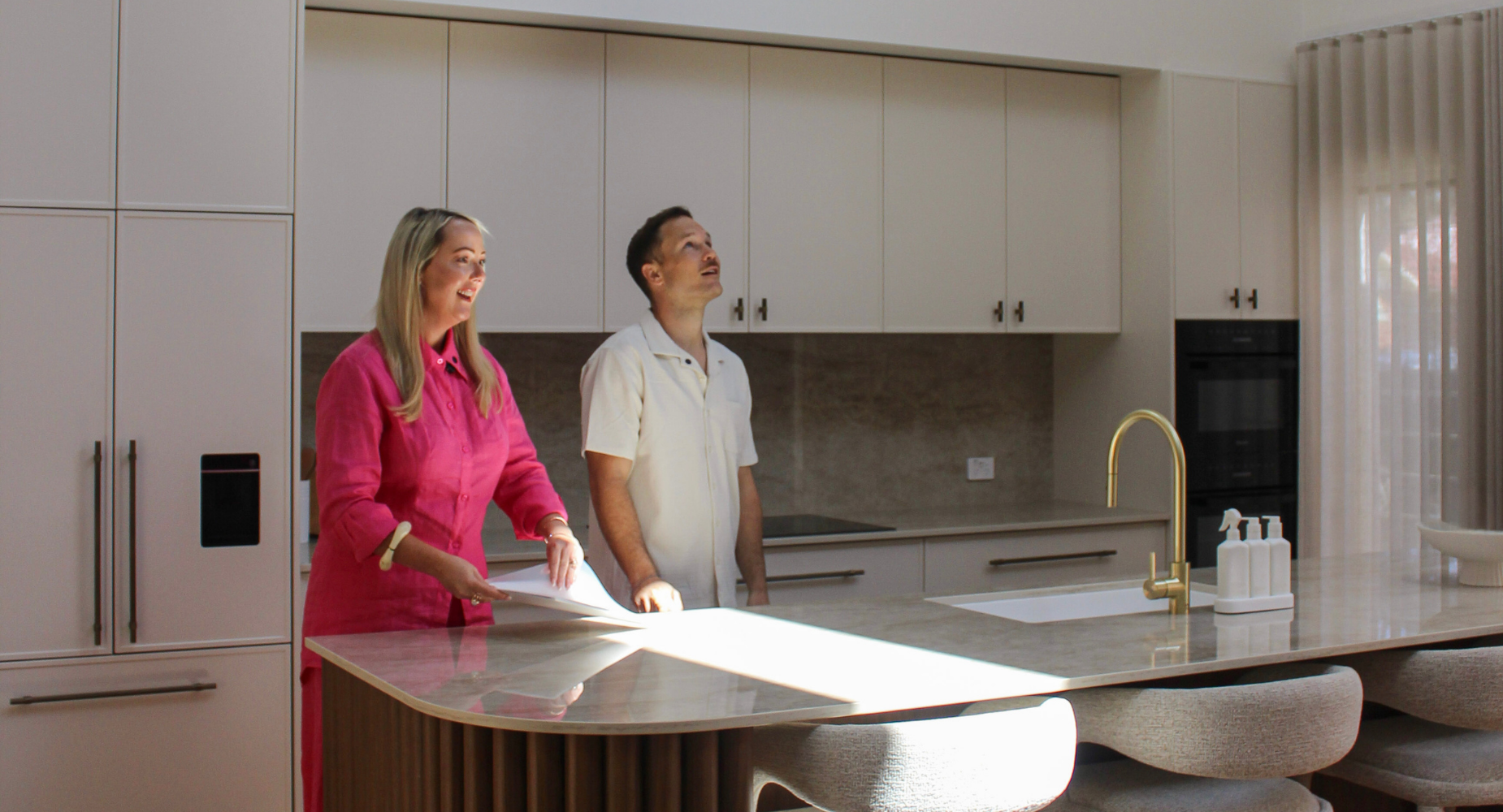 Two people, a woman in a bright pink shirt and a man in a cream shirt, standing in a modern kitchen with white cabinets and a beige countertop, engaging in conversation.