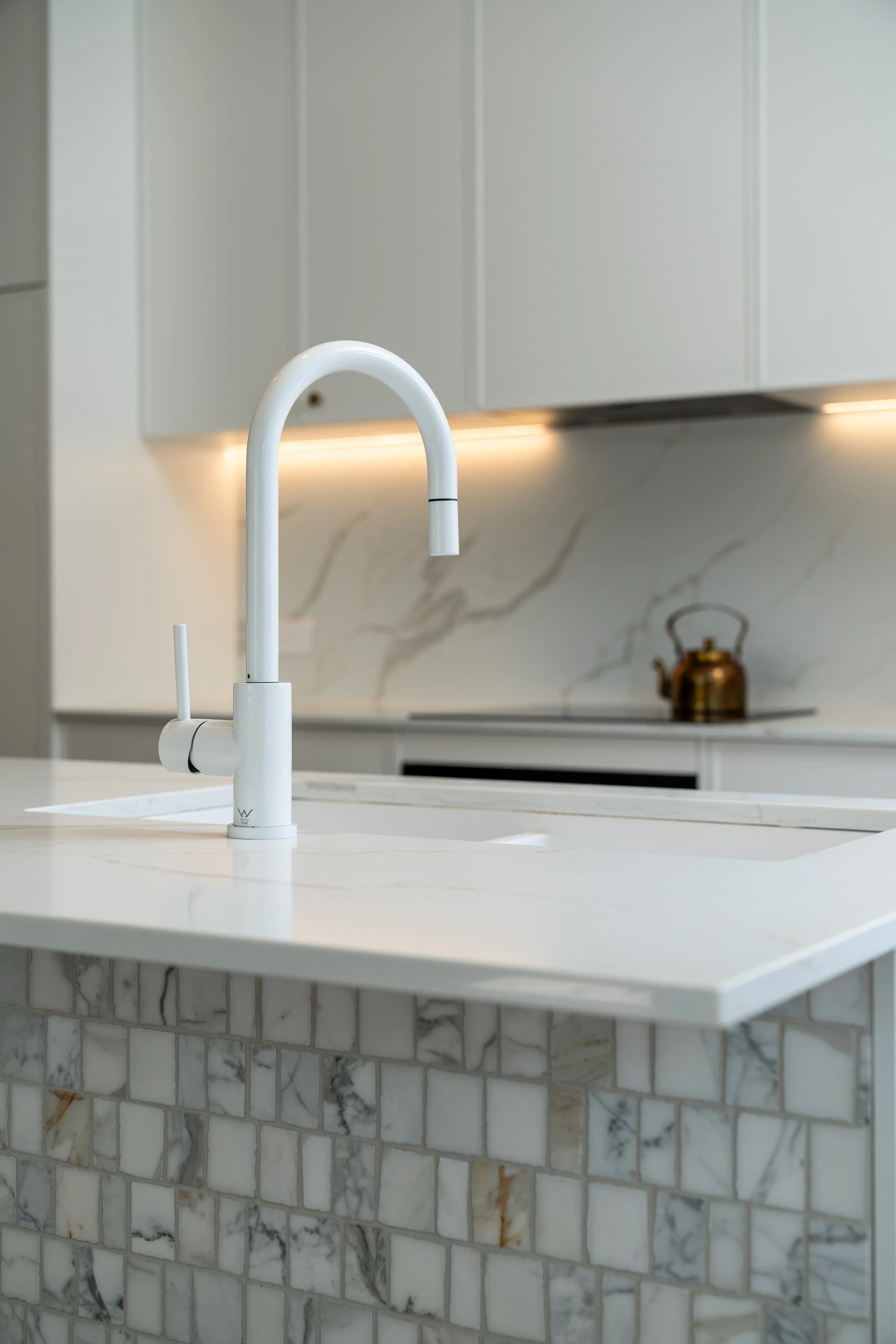 Modern kitchen with white marble countertop, white backsplash, and a white faucet over a sink. In the background, there is a copper kettle on the stovetop and white cabinets with ambient lighting.