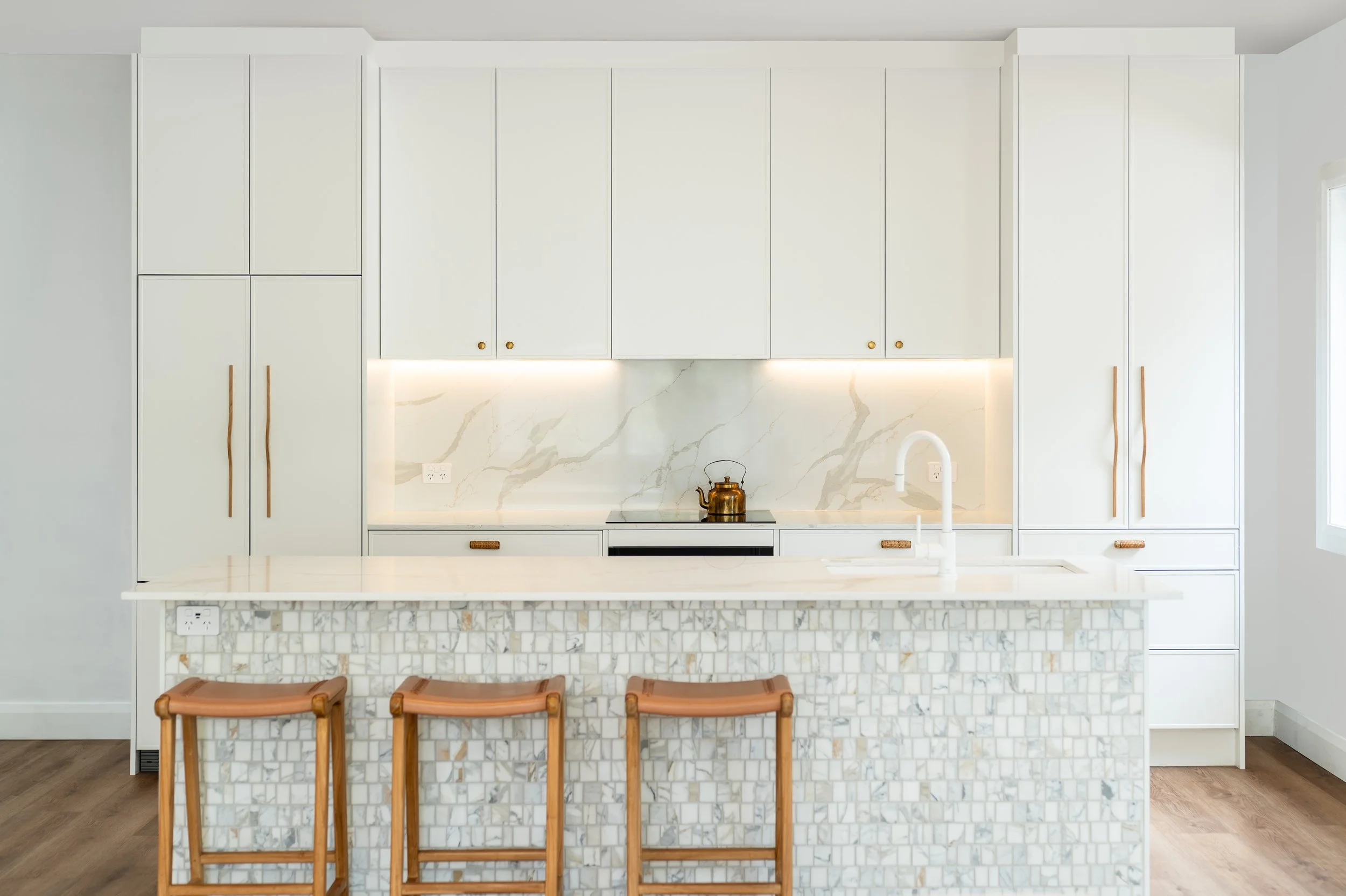 Modern kitchen with white cabinets, marble backsplash, marble countertop, a brass teapot on the stove, and three wooden stools at the island.