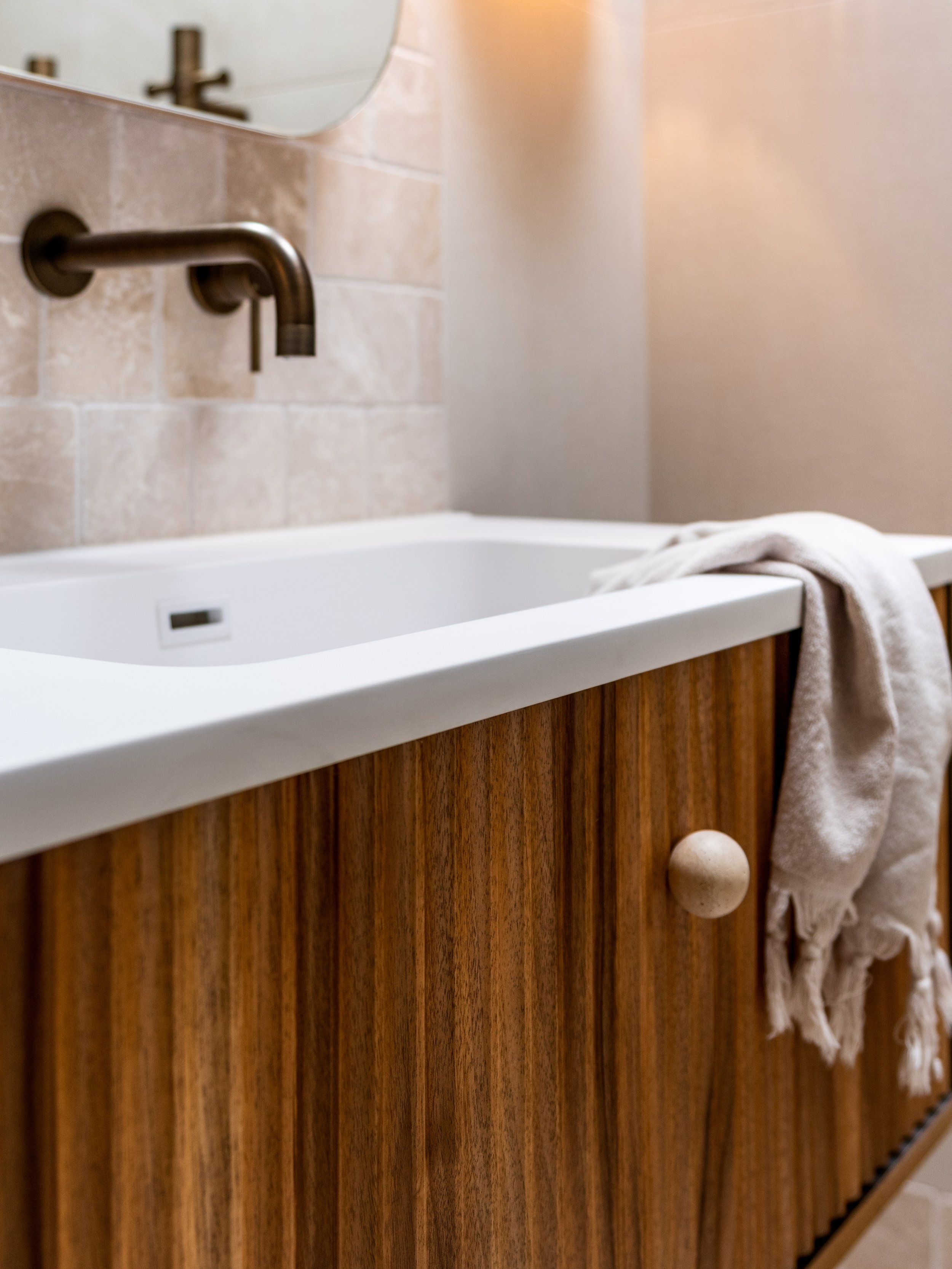 A bathroom with a white bathtub, a brass wall-mounted faucet, and a wooden cabinet with a round handle. A beige towel is draped over the edge of the tub.
