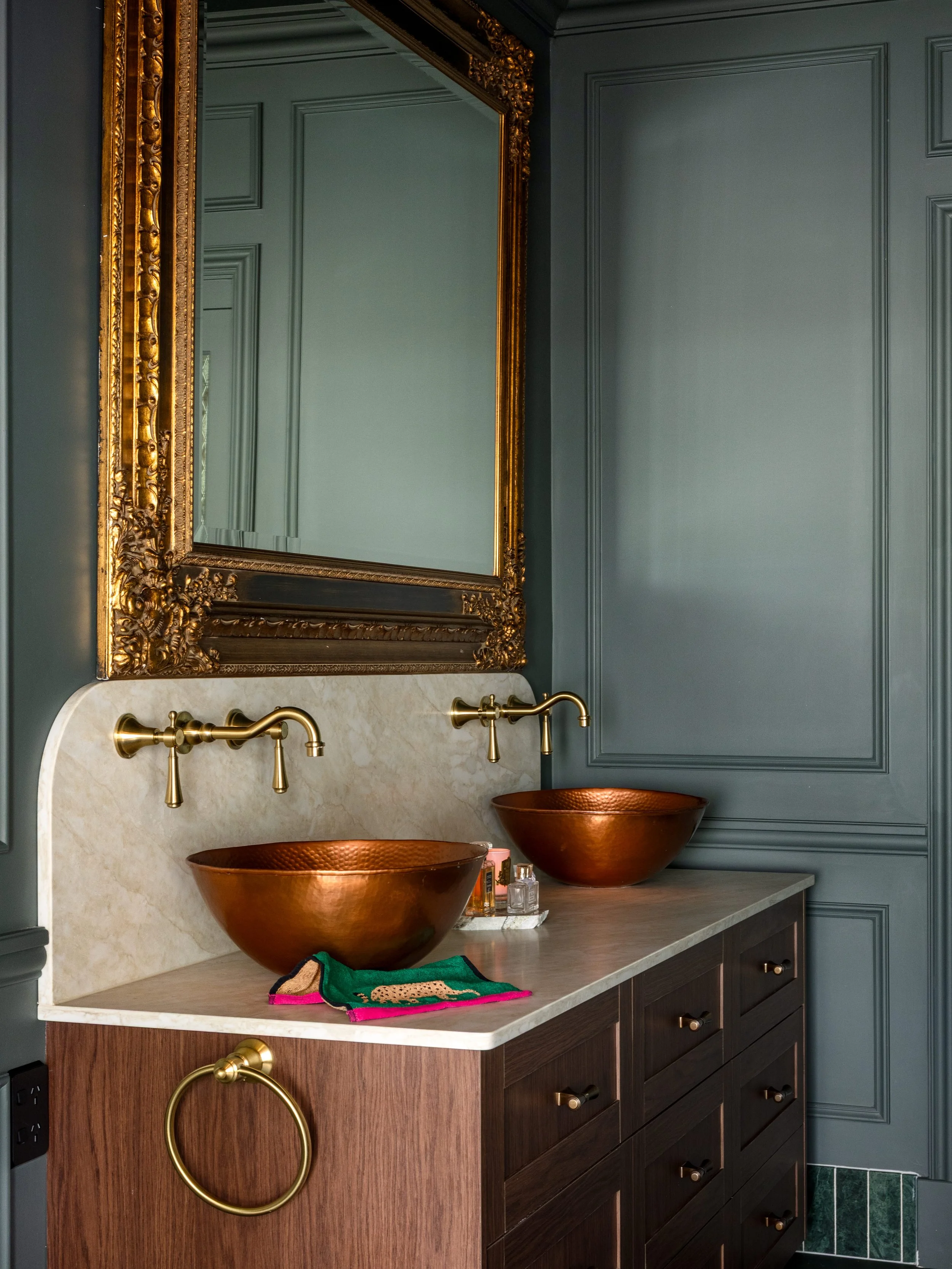 A luxurious bathroom vanity with dark wood cabinetry, a marble countertop, and copper vessel sinks. A large ornate gold-framed mirror hangs above the sinks. The wall behind is painted in a deep green/teal color with decorative molding, and there are 