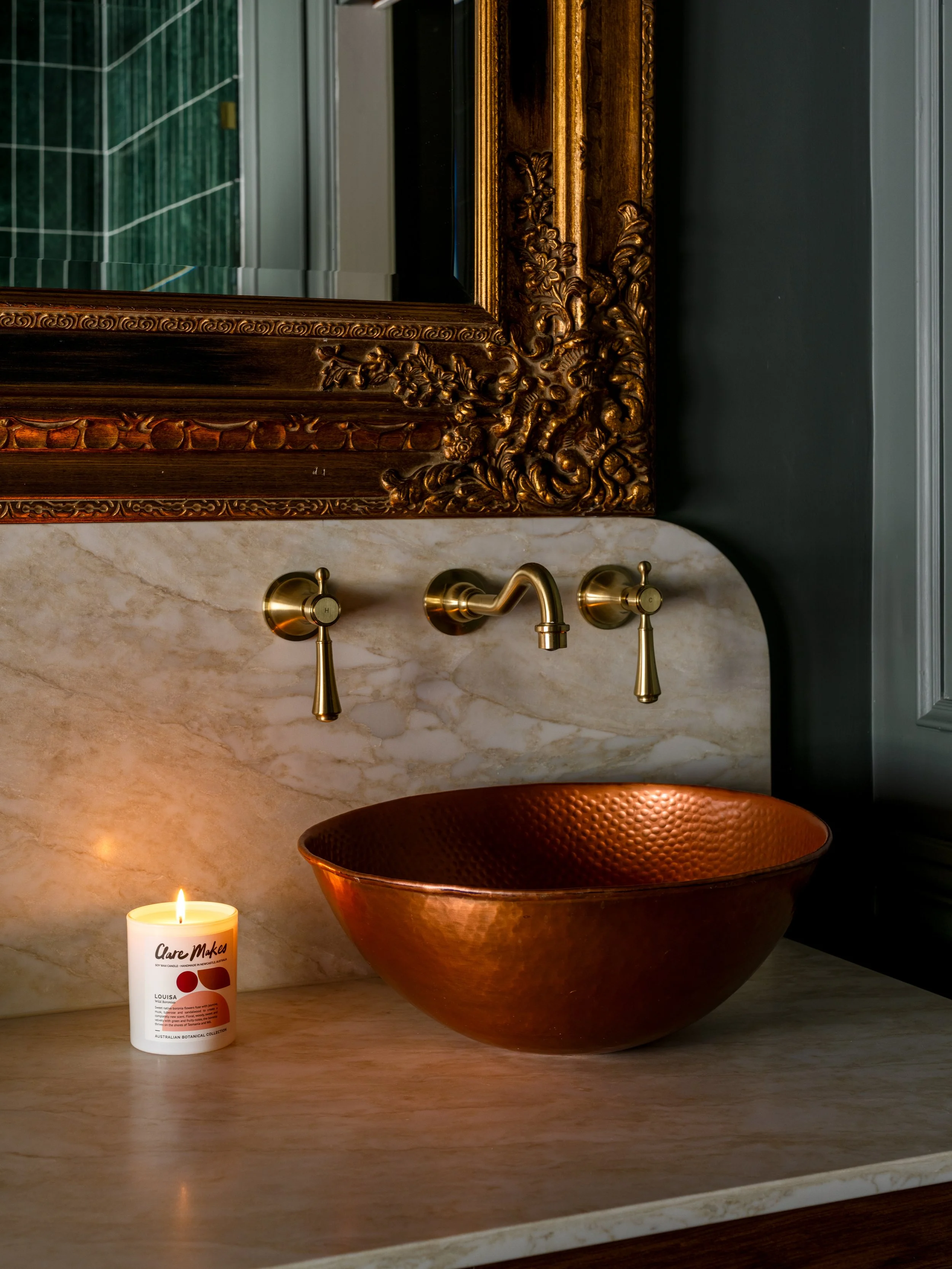 A bathroom sink area featuring a copper vessel sink, a brass faucet, a lit candle labeled 'Care Makes,' a decorative mirror with an ornate wooden frame, gray and dark green walls, and a candle on a marble countertop.