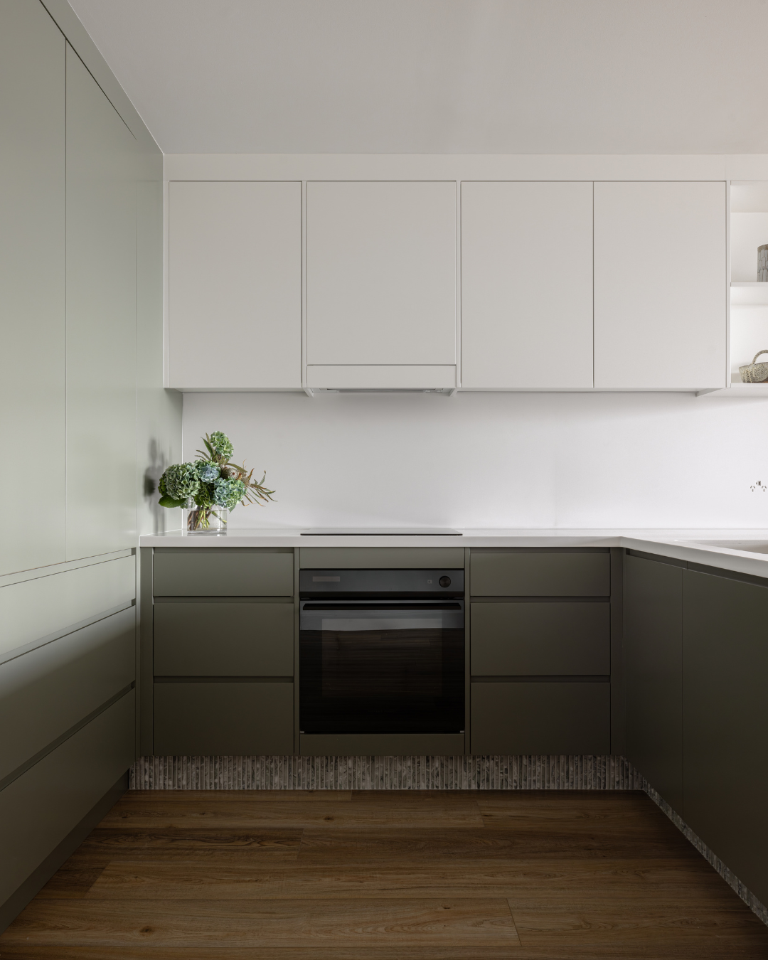 Modern minimalist kitchen with white upper cabinets, dark lower cabinets, a black oven, wooden floor, and a small bouquet of flowers on the counter.