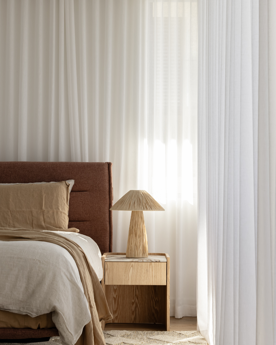 A bedroom featuring a bed with beige linens and pillows, a wooden nightstand with a matching lamp, and white curtains allowing natural light to fill the room.