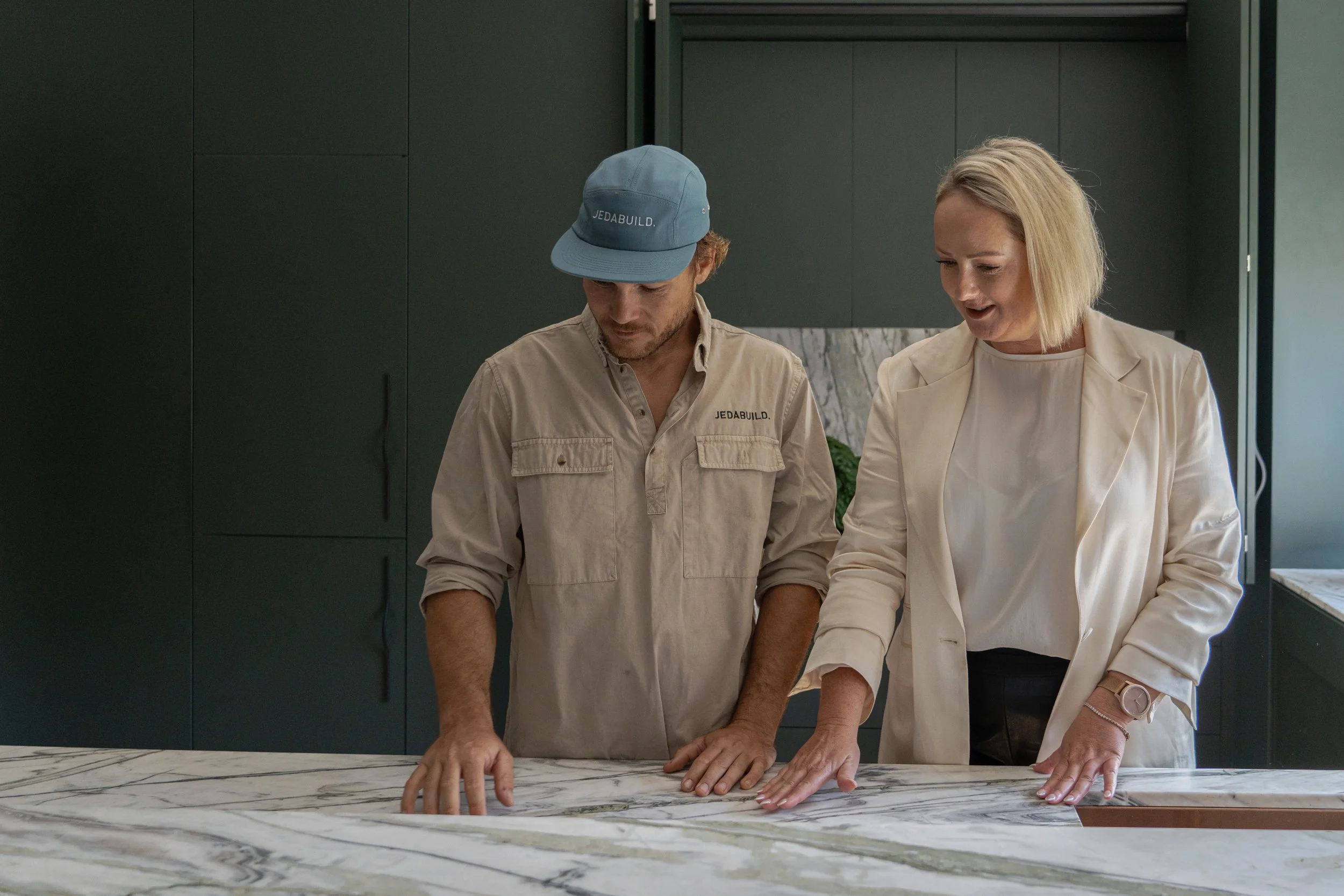 Two people, a man wearing a blue cap and beige work shirt and a woman in a cream blazer, are inspecting a large marble surface with their hands on it in a modern kitchen.