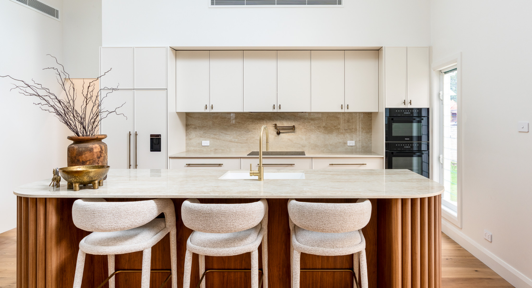 Modern kitchen with a marble island, beige cabinetry, and black appliances. Decor includes a large vase with branches and a gold bowl on the island, with barstools underneath.